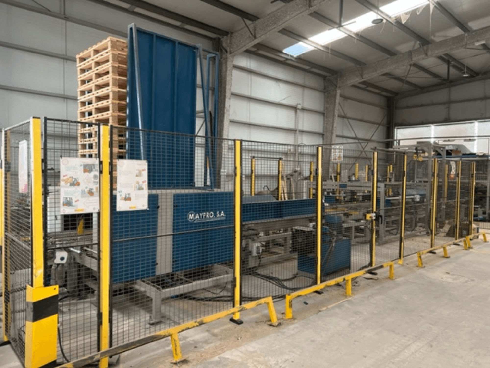 Maypro S.A. rūpnieciskā palešu iepakošanas iekārta noliktavā, ar drošības sietu un vadības ierīcēm, skats no sāniem.