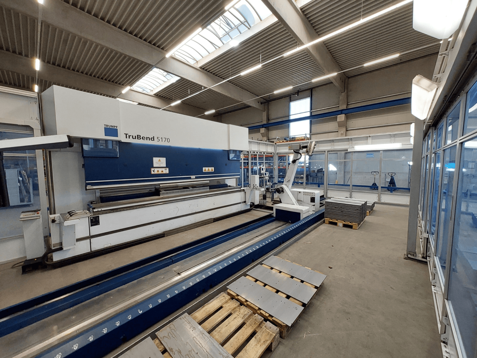 TRUMPF TruBend 5170 preses bremze darbnīcā, skats no sāniem, redzams liekšanas instruments un uz paliktņiem sakrautas metāla loksnes.