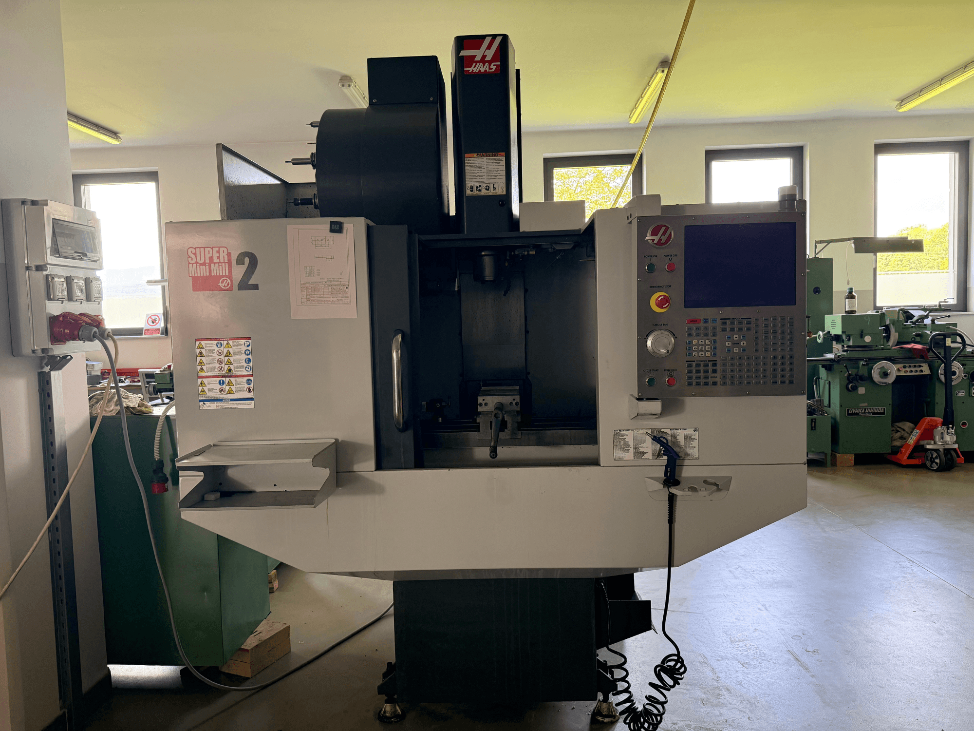 Haas Mini Mill 2 vertikālais apstrādes centrs priekšpusē, kurā redzams vadības panelis un instrumentu zona ar brīdinājuma uzrakstiem.