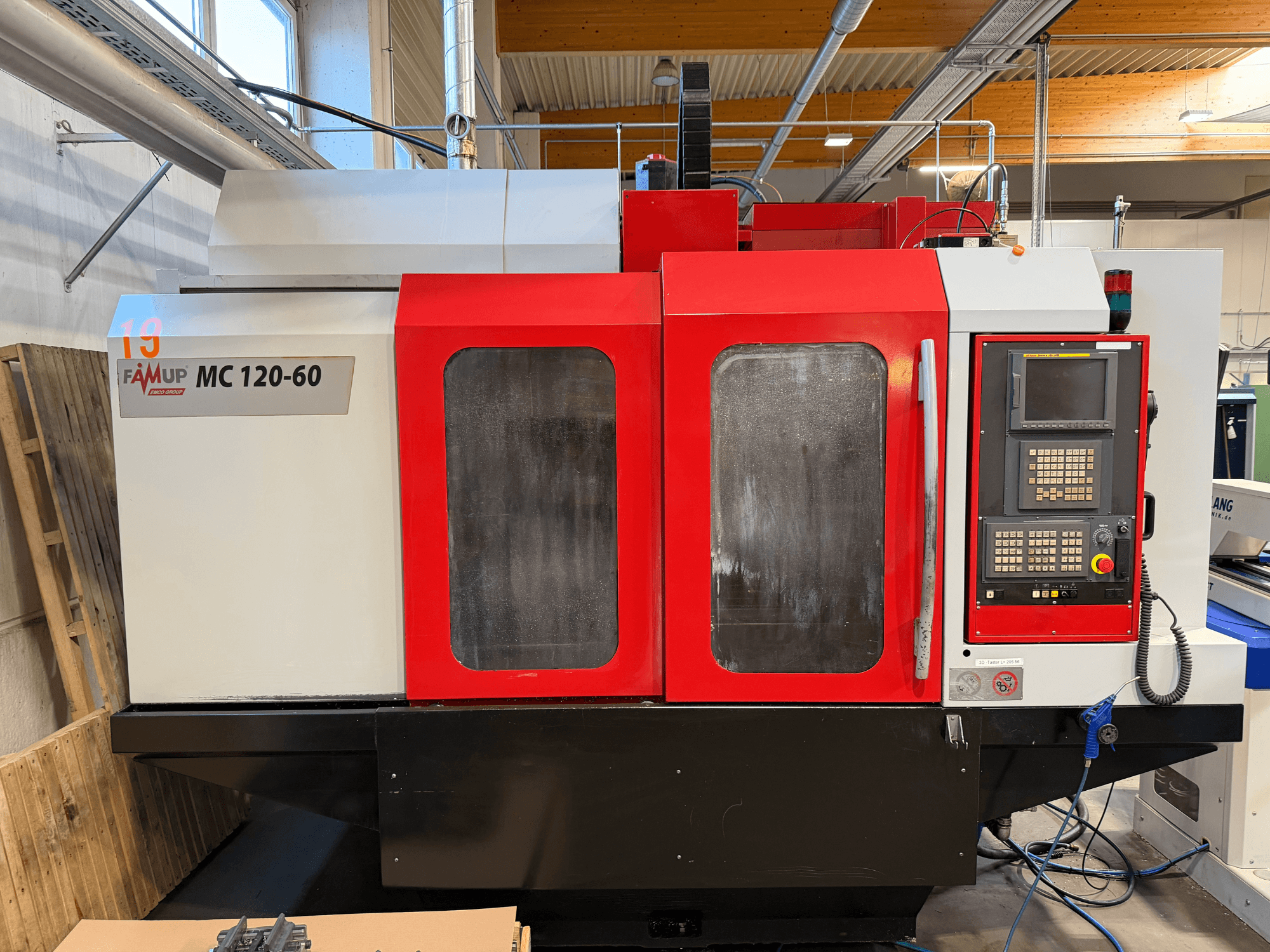 EMCO MC 120-60 vertikālā apstrādes centra priekšējais skats ar sarkanbaltsarkano korpusu, vadības paneli un displeja ekrānu.