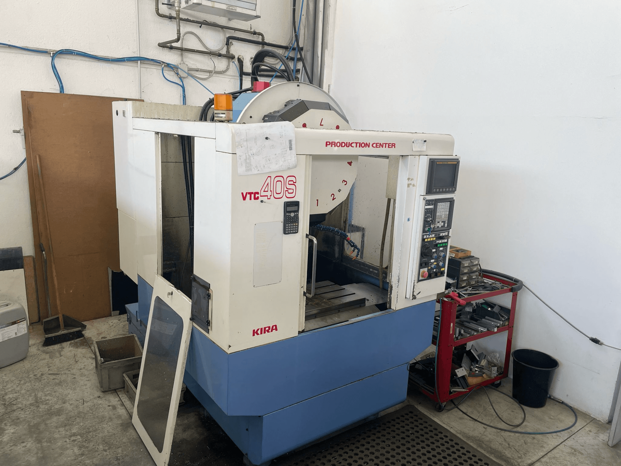 Apstrādes centrs KIRA VTC 40S, skats no sāniem, ar vadības paneli, atvērtām durvīm un instrumentiem uz uzglabāšanas ratiņiem blakus.
