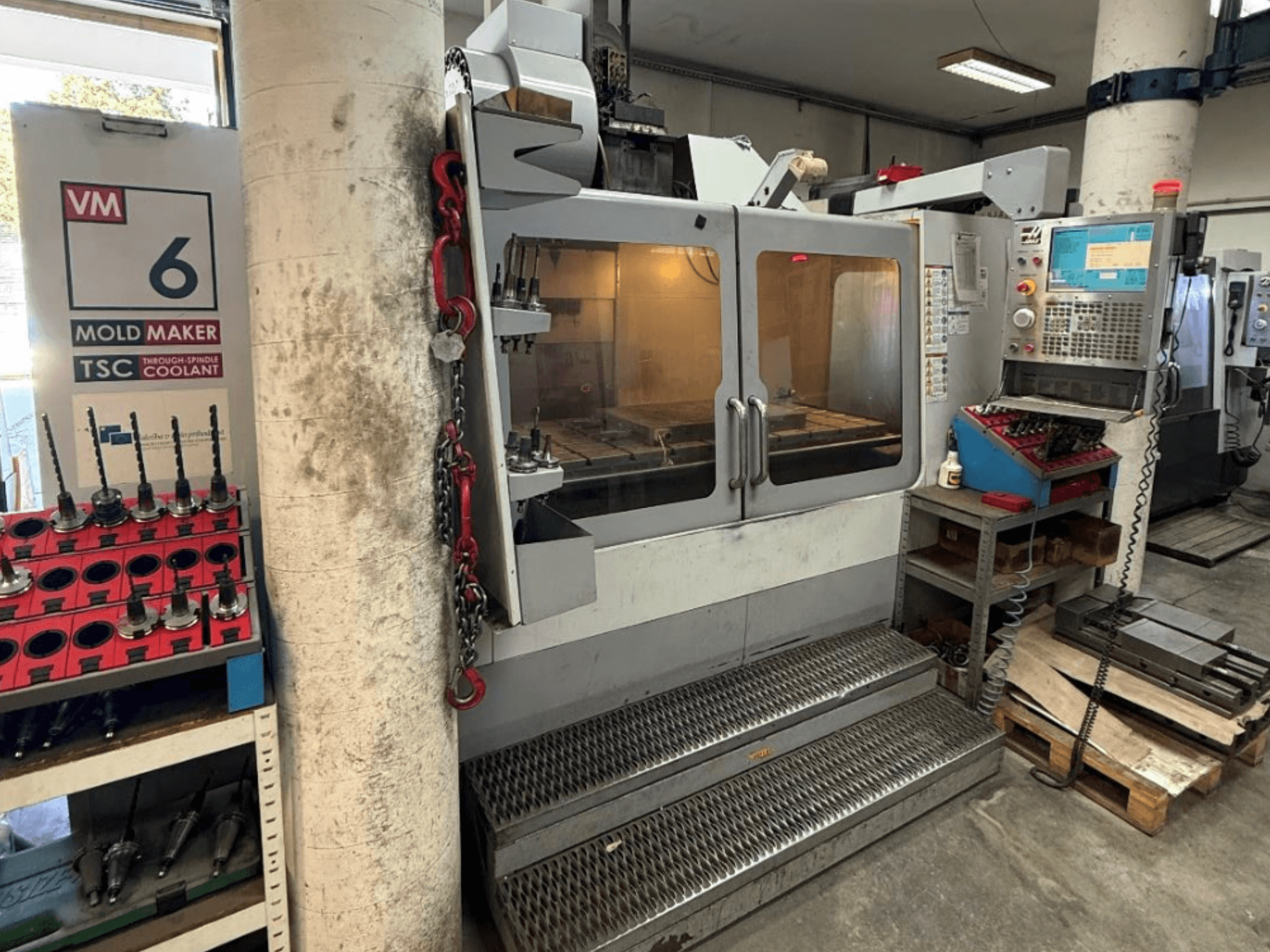 HAAS VM-6 HE vertikālais apstrādes centrs, skats no priekšpuses, kurā redzamas vadības ierīces, dzesēšanas sistēma un instrumentu glabātava.