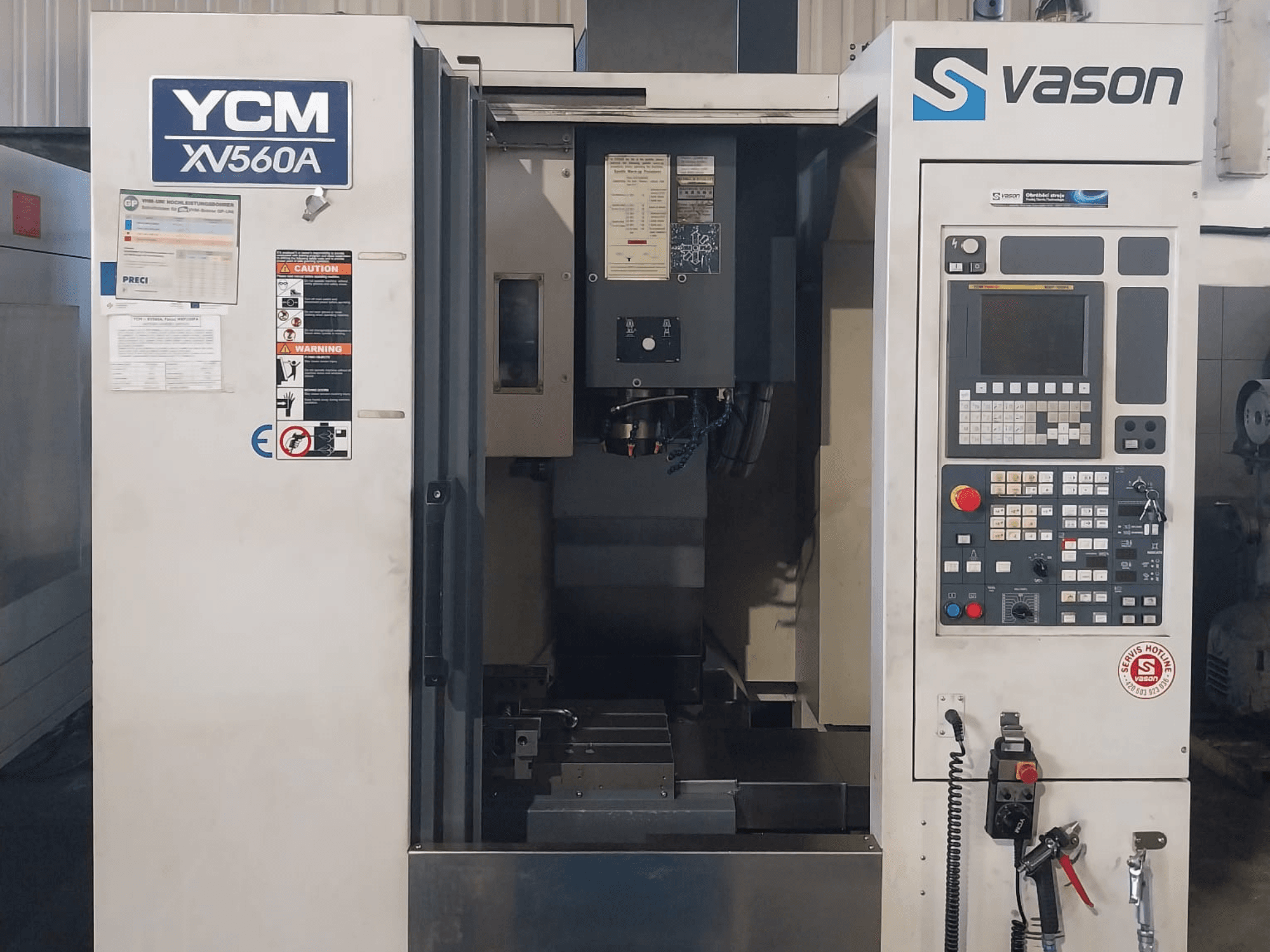 YCM XV 560A vertikālā apstrādes centra priekšējais skats ar vadības paneli un darba galdu, uz kura redzami apstrādes komponenti.