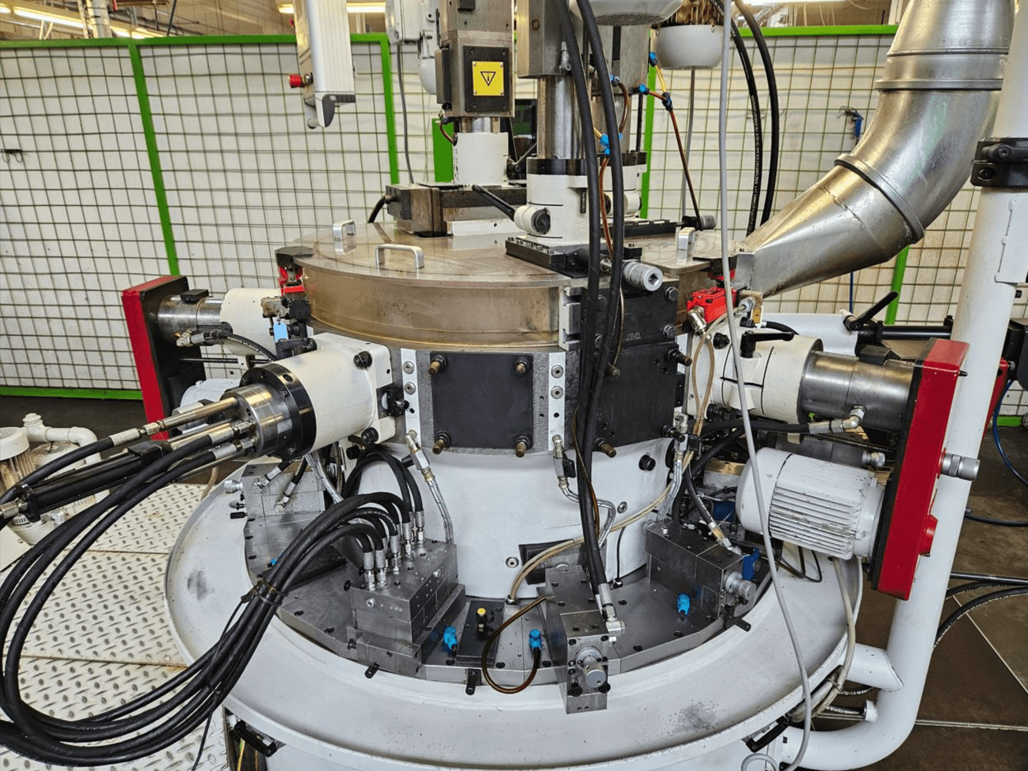 Rūpnieciskas mašīnas tuvplāns ar hidrauliskajām šļūtenēm, metāla komponentiem un rotējošu pamatni. Nav redzams zīmols.