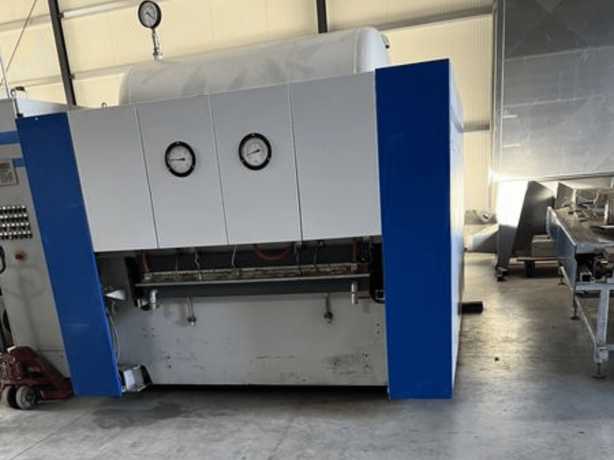 Zili balta rūpnieciska iekārta ar diviem manometriem, skats no priekšpuses, ar vadības paneli un blakus esošajām iekārtām.