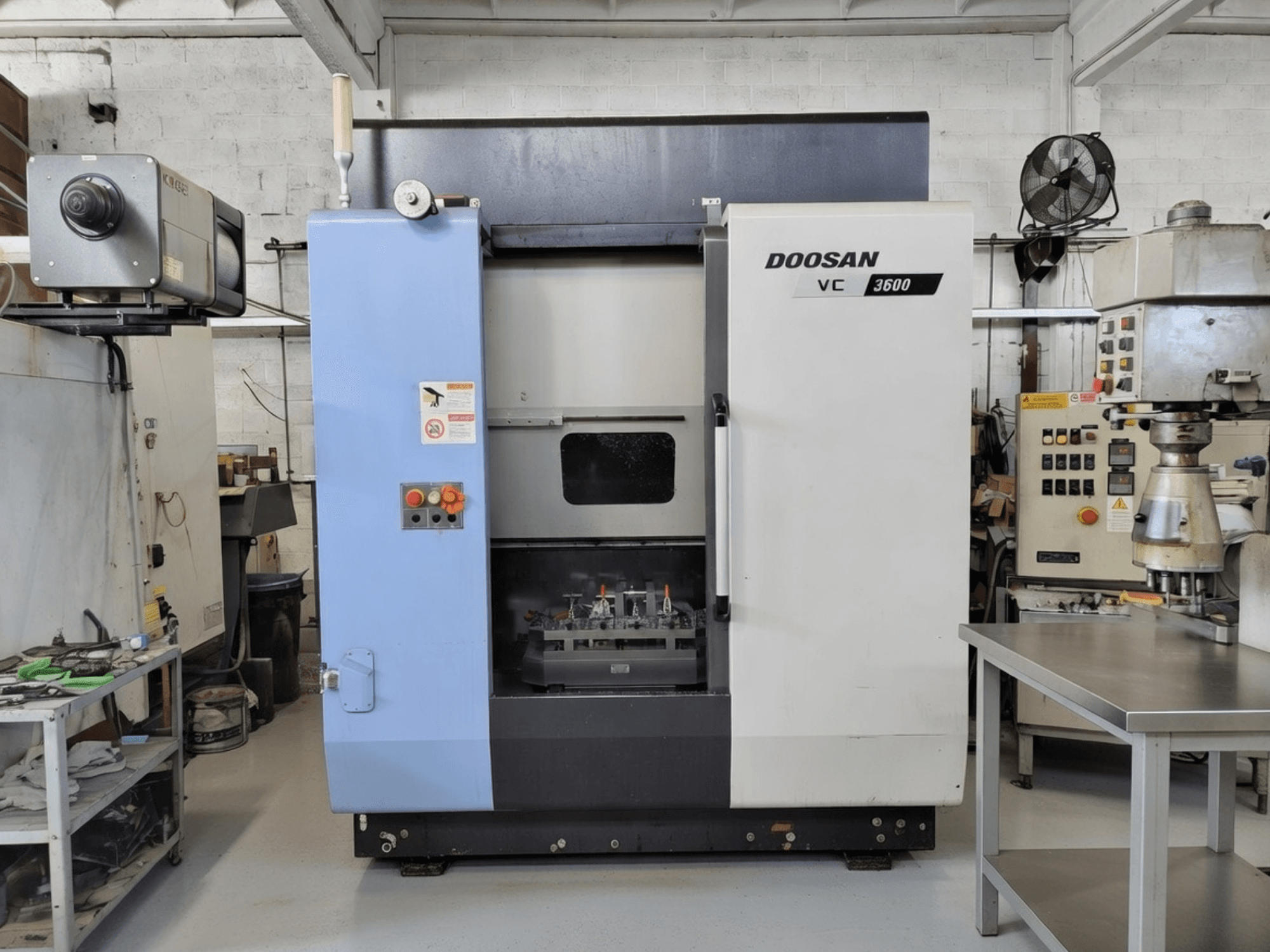 DOOSAN VC 3600 vertikālais apstrādes centrs priekšpusē, ar vadības paneli un atvērtām durvīm, kas atklāj darba vietu.
