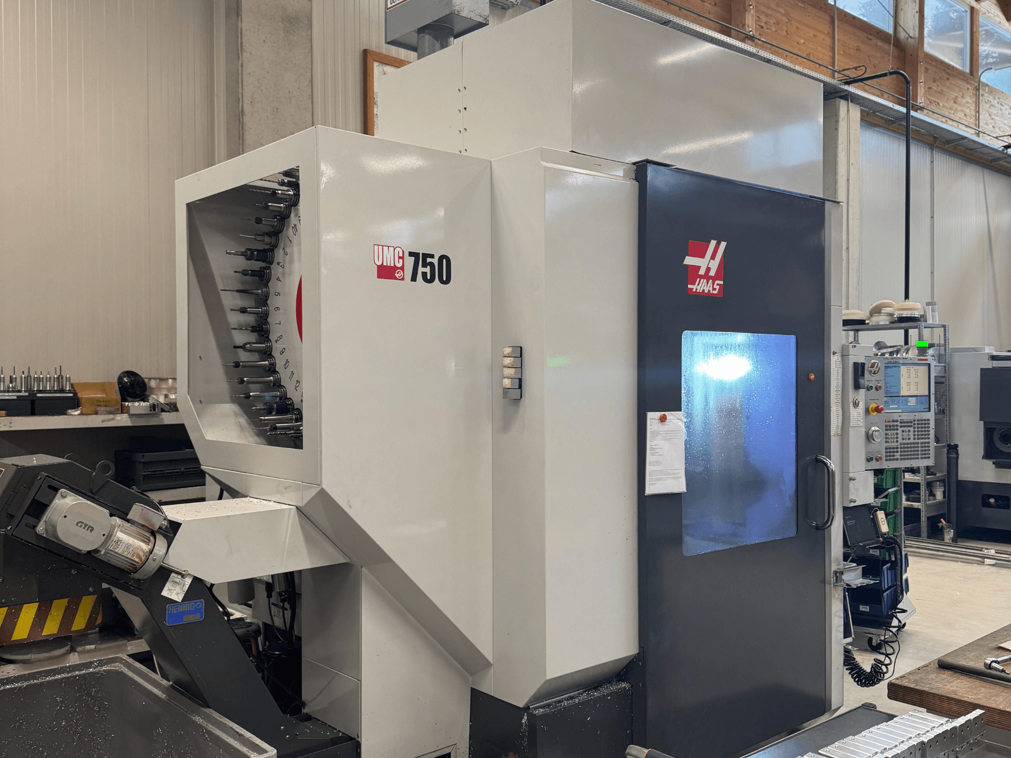 Haas UMC 750 apstrādes centrs, priekšējais skats, kurā redzams instrumentu turētājs un vadības panelis darbnīcā.