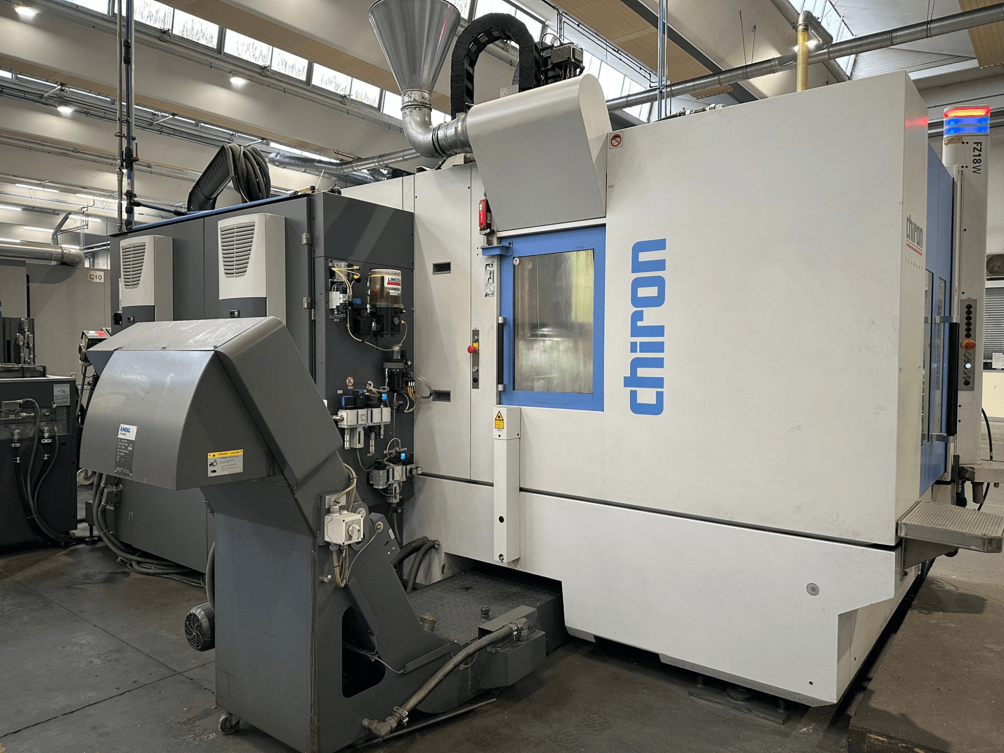 Chiron CNC mašīna pelēkā un baltā krāsā, skats no priekšpuses, ar vadības ierīcēm, caurulēm un putekļu nosūces pārsegu.