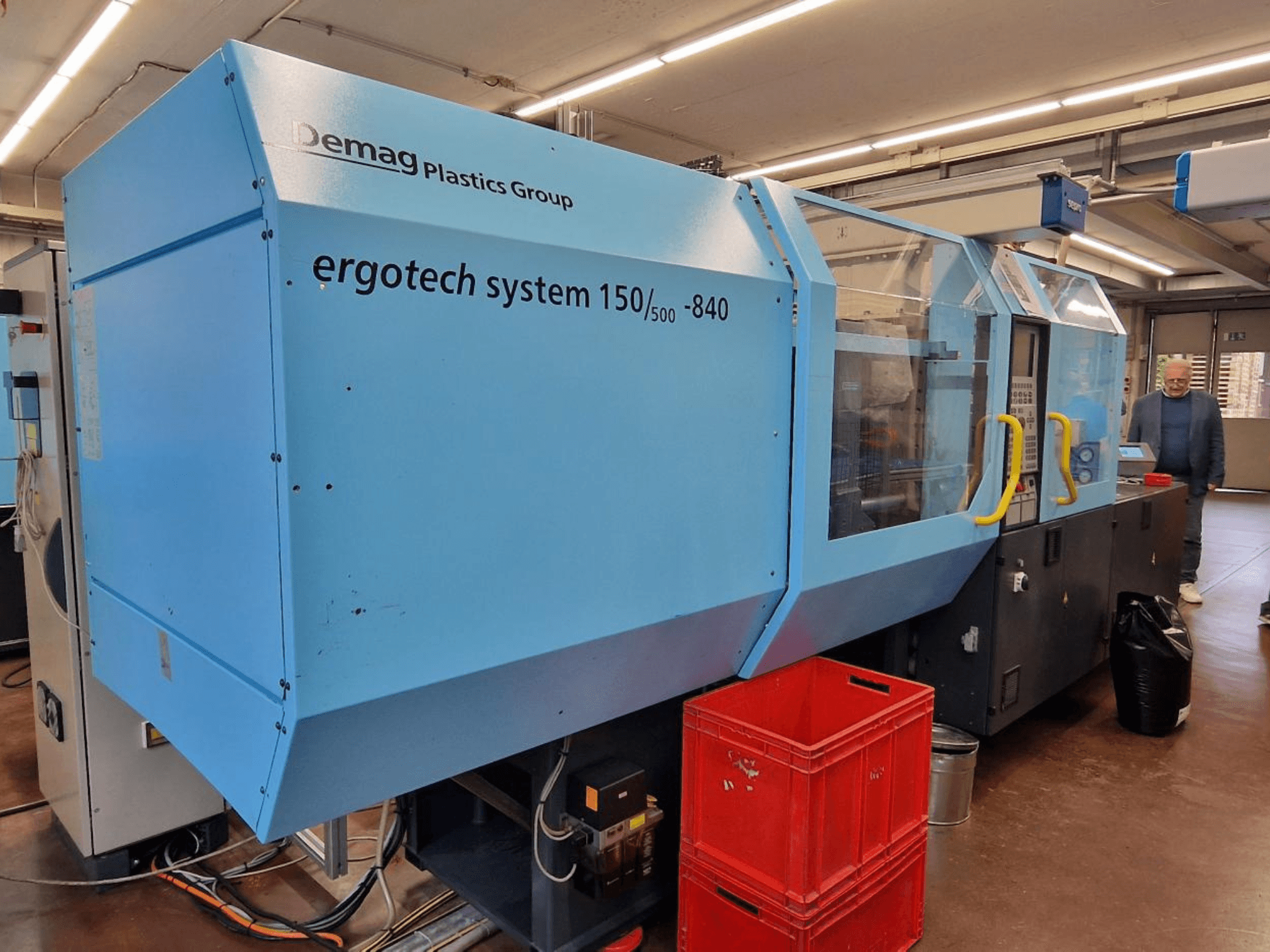 Demag Ergotech System 150/500-840 injekcijas formēšanas iekārta zilā krāsā, skats no sāniem, ar kastēm un operatoru fonā.