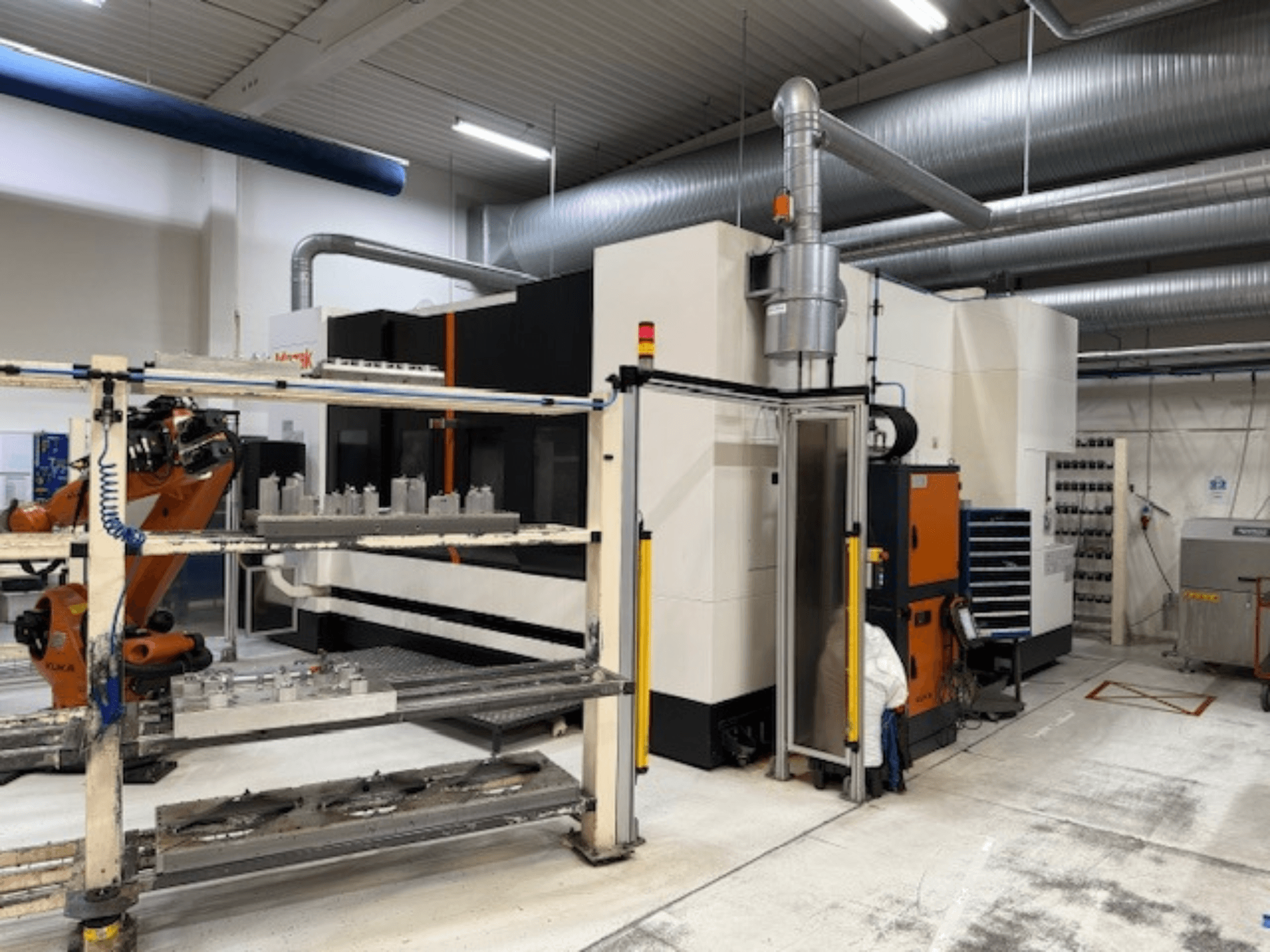 Rūpnieciskais CNC darbagalds ar robotizētu roku un drošības barjerām, ar skatu uz iekārtu no priekšpuses, gaisīgā darba telpā.
