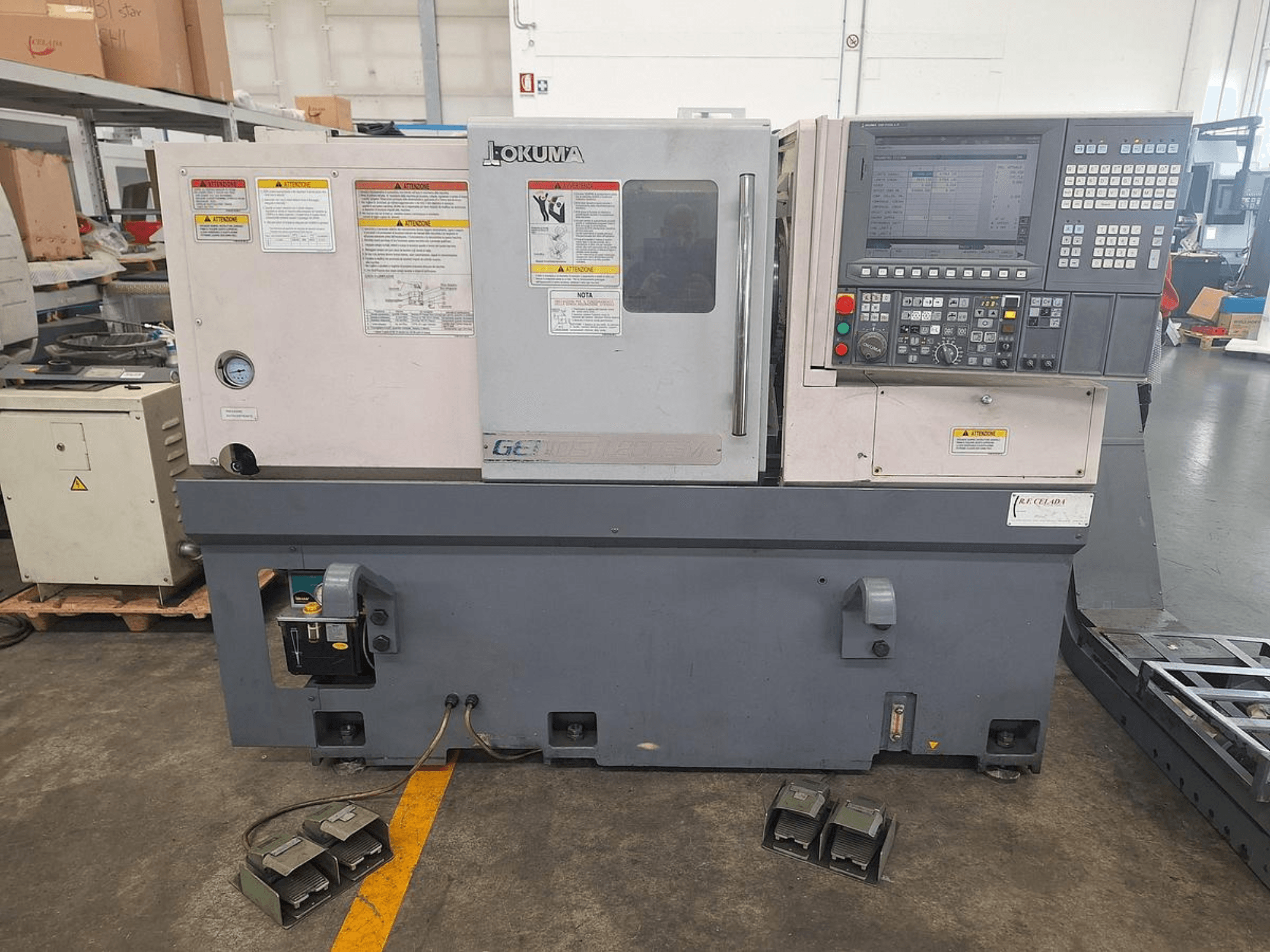 Okuma GENOS L200E-M horizontālās virpošanas iekārta no sāniem; vadības panelis, drošības brīdinājumi un pedāļi.