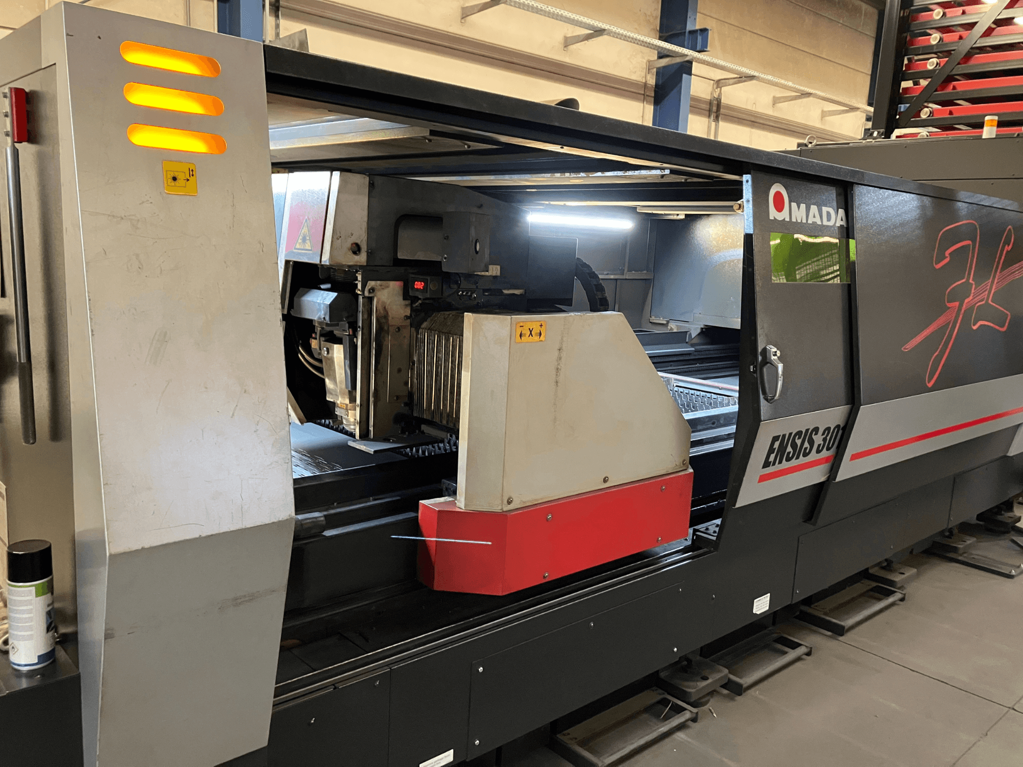 AMADA ENSIS 3015 AJ šķiedru lāzera griešanas iekārta, skats no priekšpuses, redzama griešanas galva un vadības ierīces metāla rāmī.