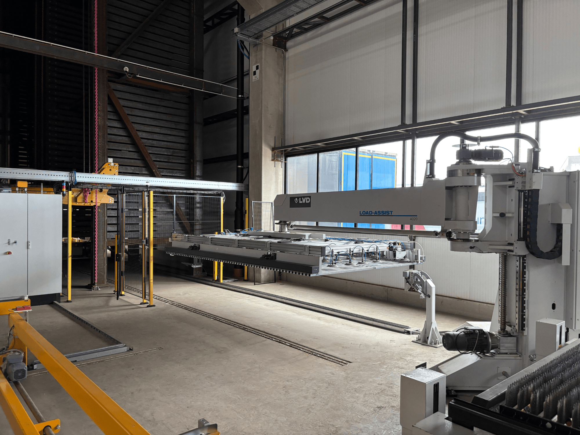 LVD Load-Assist 4020 automatizācijas iekārta priekšpusē ar robotizētu roku un iekraušanas sistēmu, kas redzama lielā darba telpā.