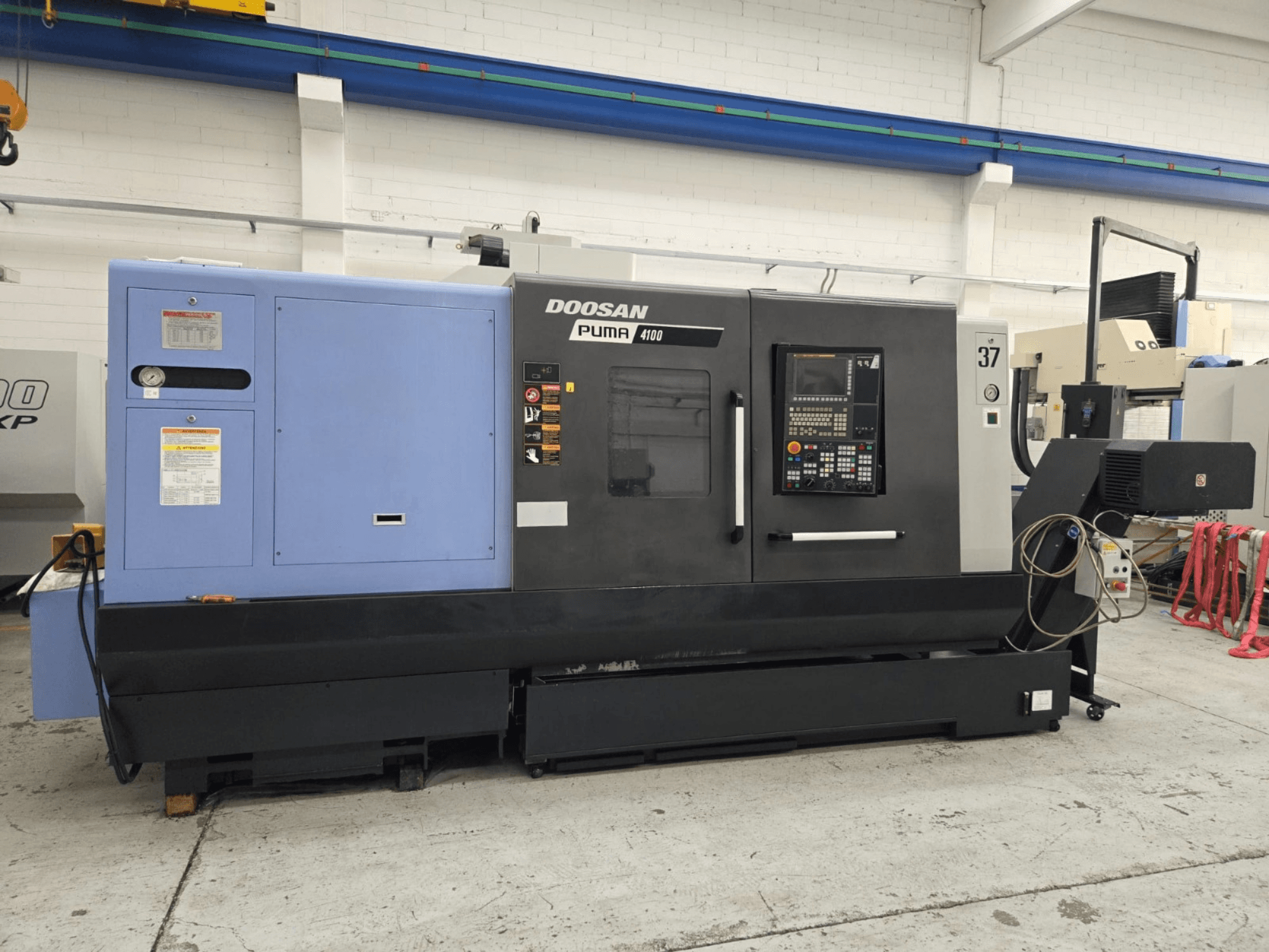 Doosan Puma 4100 CNC virpa, skats no priekšpuses; vadības panelis, zilais barošanas bloks un detalizētas uzlīmes darbības norādījumiem.