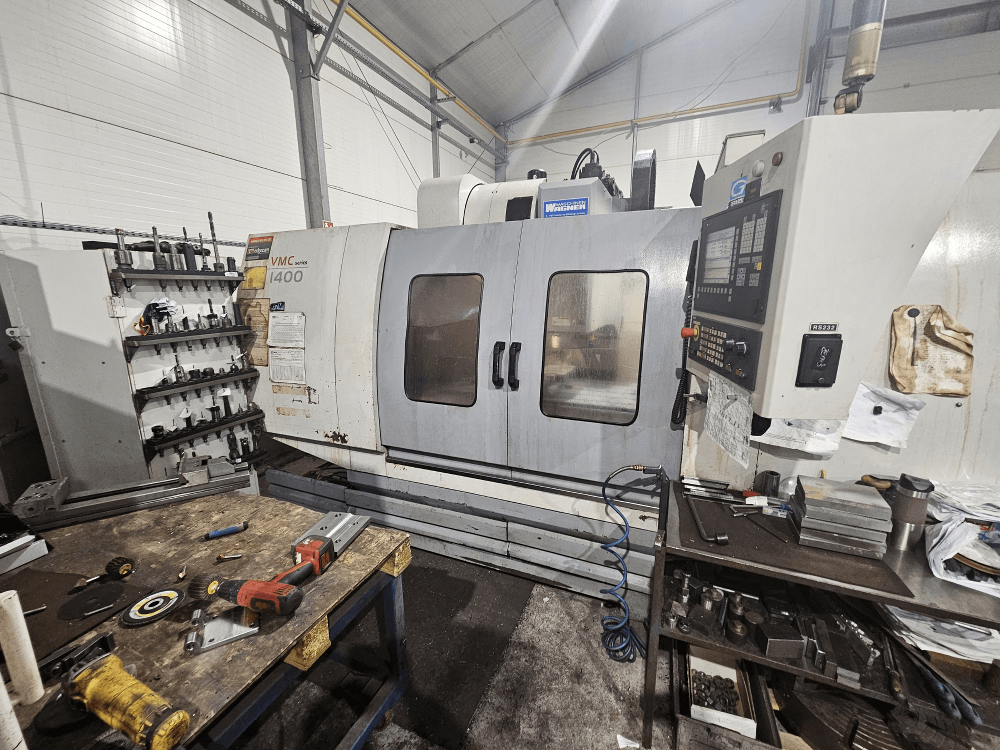 VMC 1400 CNC frēzmašīna darbnīcā, skats no priekšpuses; instrumenti un materiāli izkaisīti uz darbgalda blakus.