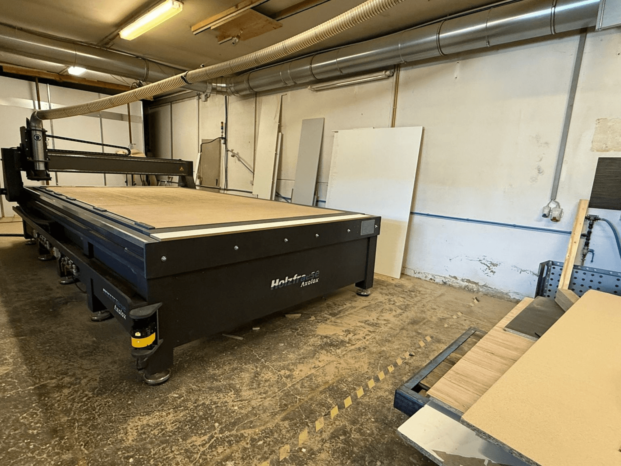 Holz-Her Axos 5 CNC frēzētājs darbnīcā, skats no priekšpuses, kur redzams darba galds, putekļu nosūces sistēma un koka paneļi blakus.