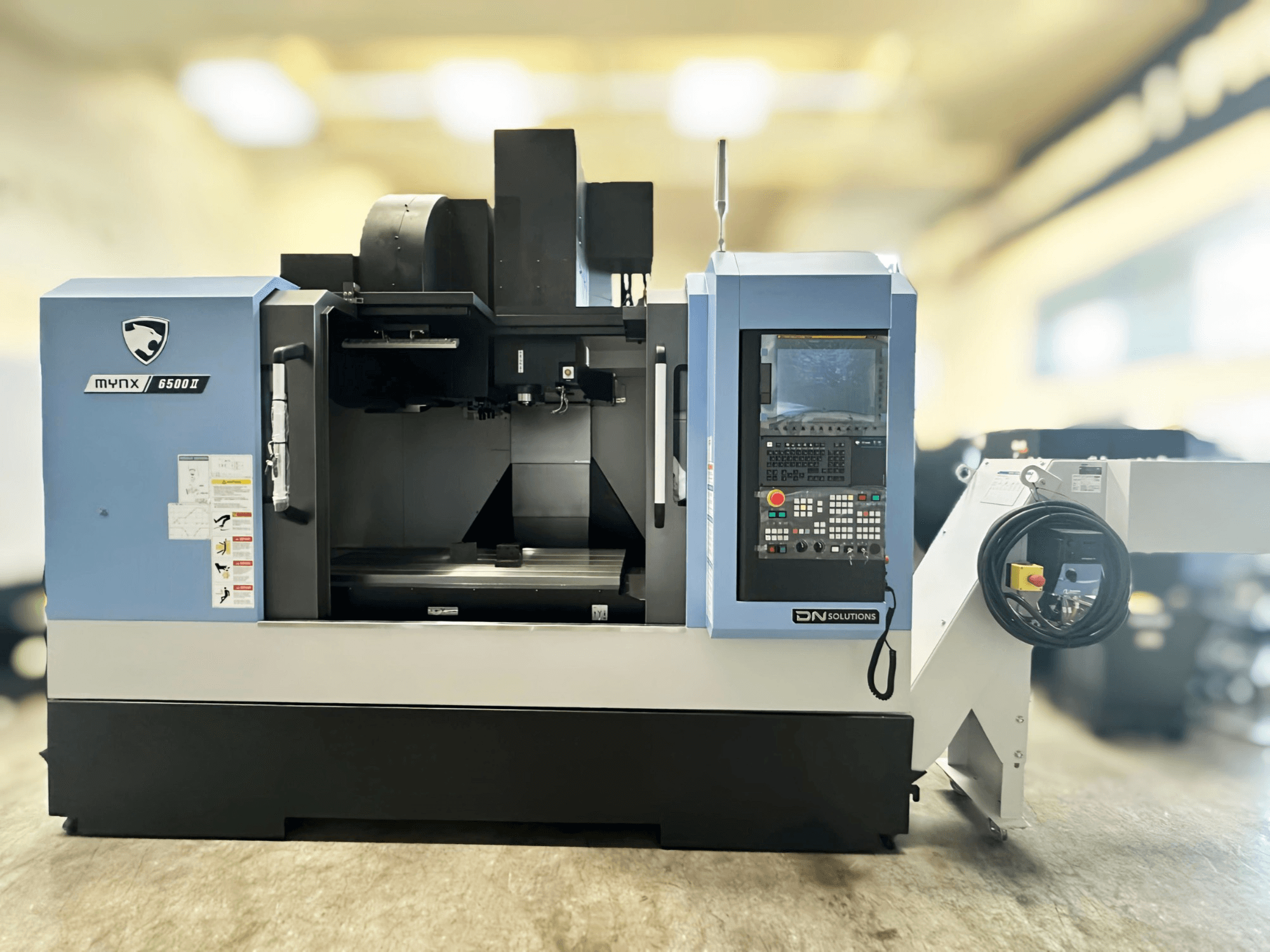 DN SOLUTIONS MYNX 6500 II vertikālais apstrādes centrs, skats no priekšpuses, kurā redzams vadības panelis un darbības saskarne.