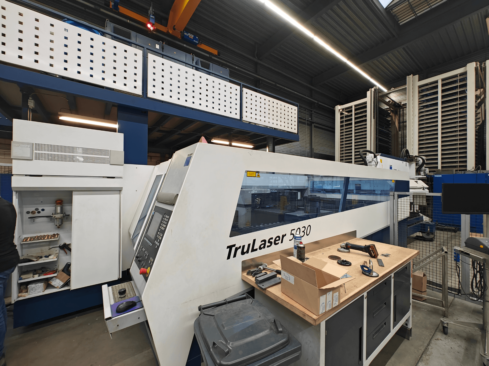 TruLaser 5030 mašīna darbnīcā, skats no priekšpuses, redzams vadības panelis, ap to ir instrumenti un aprīkojums.
