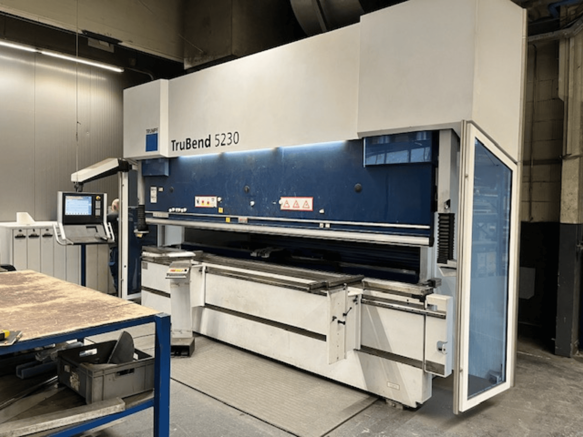 TruBend 5230 liekšanas darbagalds rūpnieciskā vidē; priekšējais skats, kurā redzams vadības panelis, drošības elementi un darba galds.