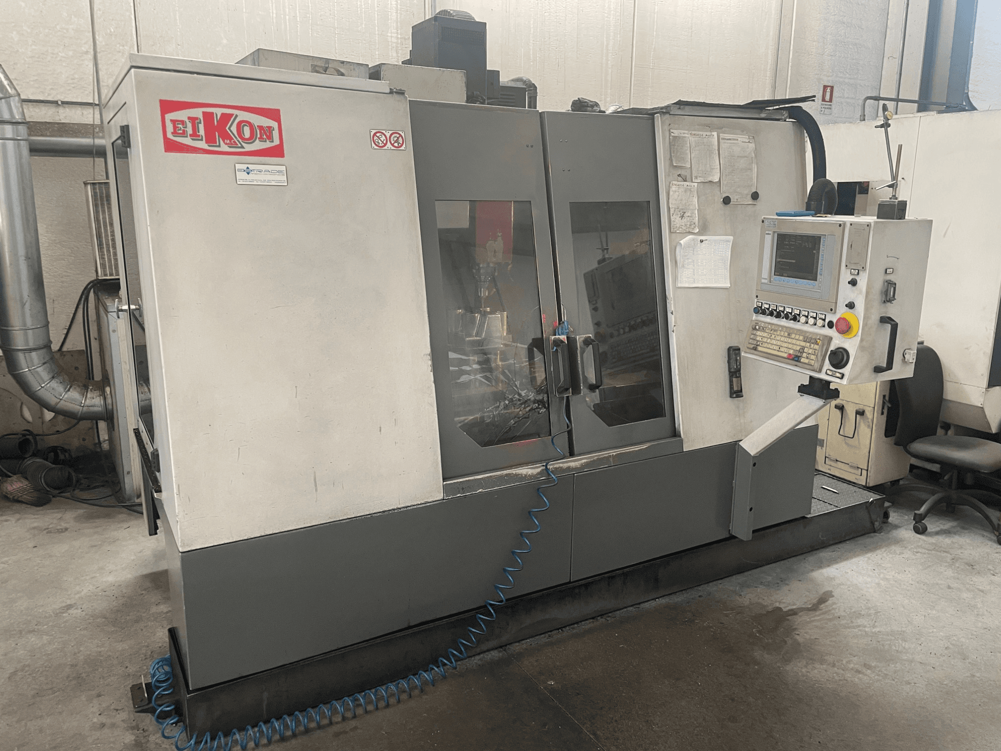 EIKON CNC mašīna no priekšpuses, ar vadības paneli, lielām stikla durvīm un zilu gaisa šļūteni uz grīdas.