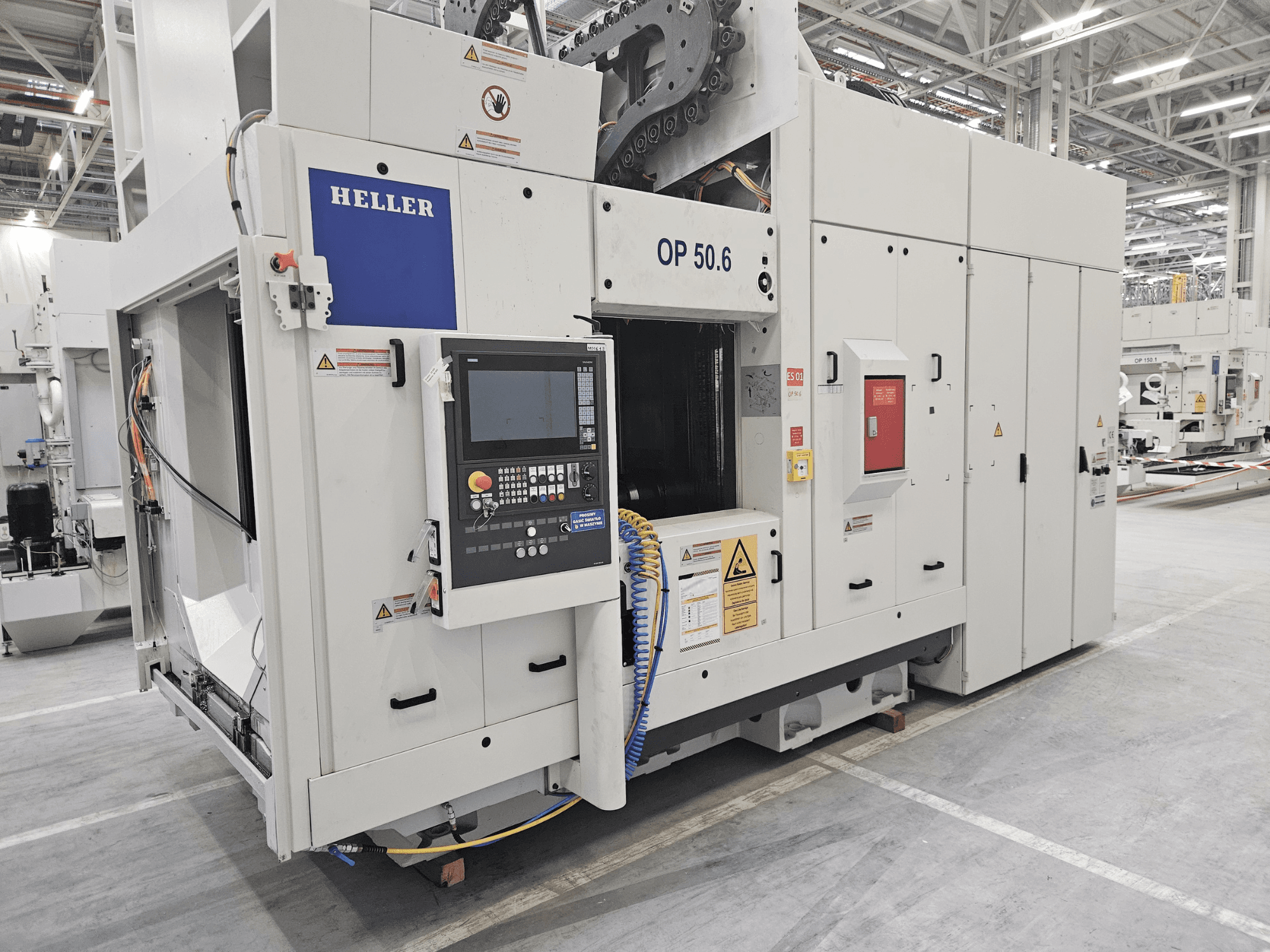 Heller OP 50.6 apstrādes centrs, skats no priekšpuses, ar vadības paneli, brīdinājuma uzlīmes un šļūtenes redzamas rūpnīcā.