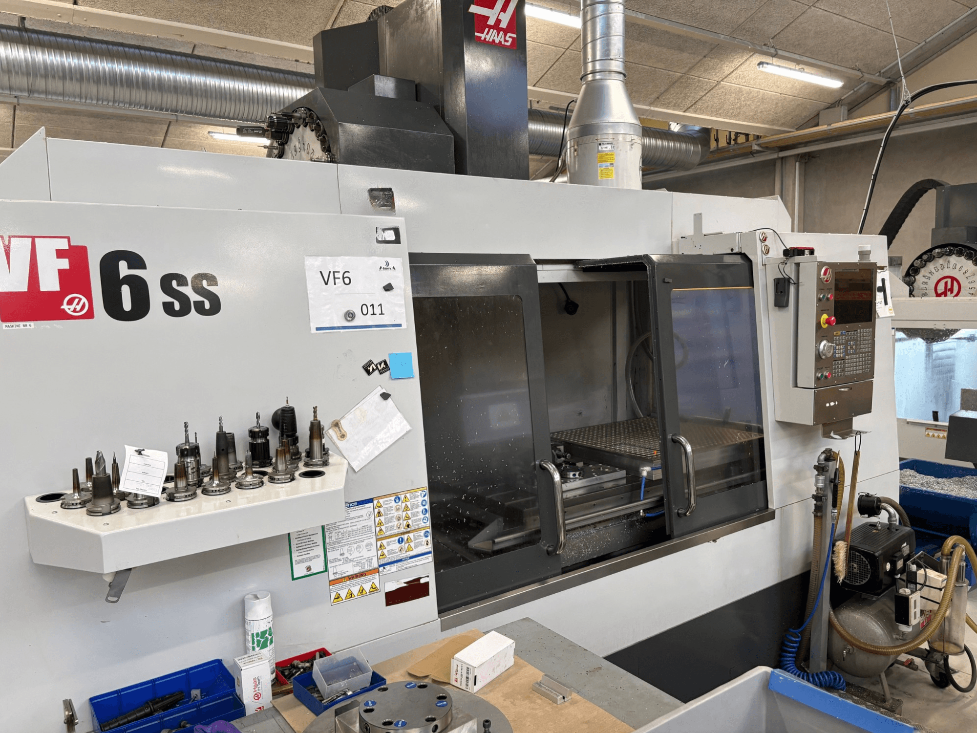 Haas VF-6 SS CNC frēzmašīna, skats no priekšpuses. Ar dažādiem instrumentiem, vadības paneli un redzamu iekšējo darba telpu.