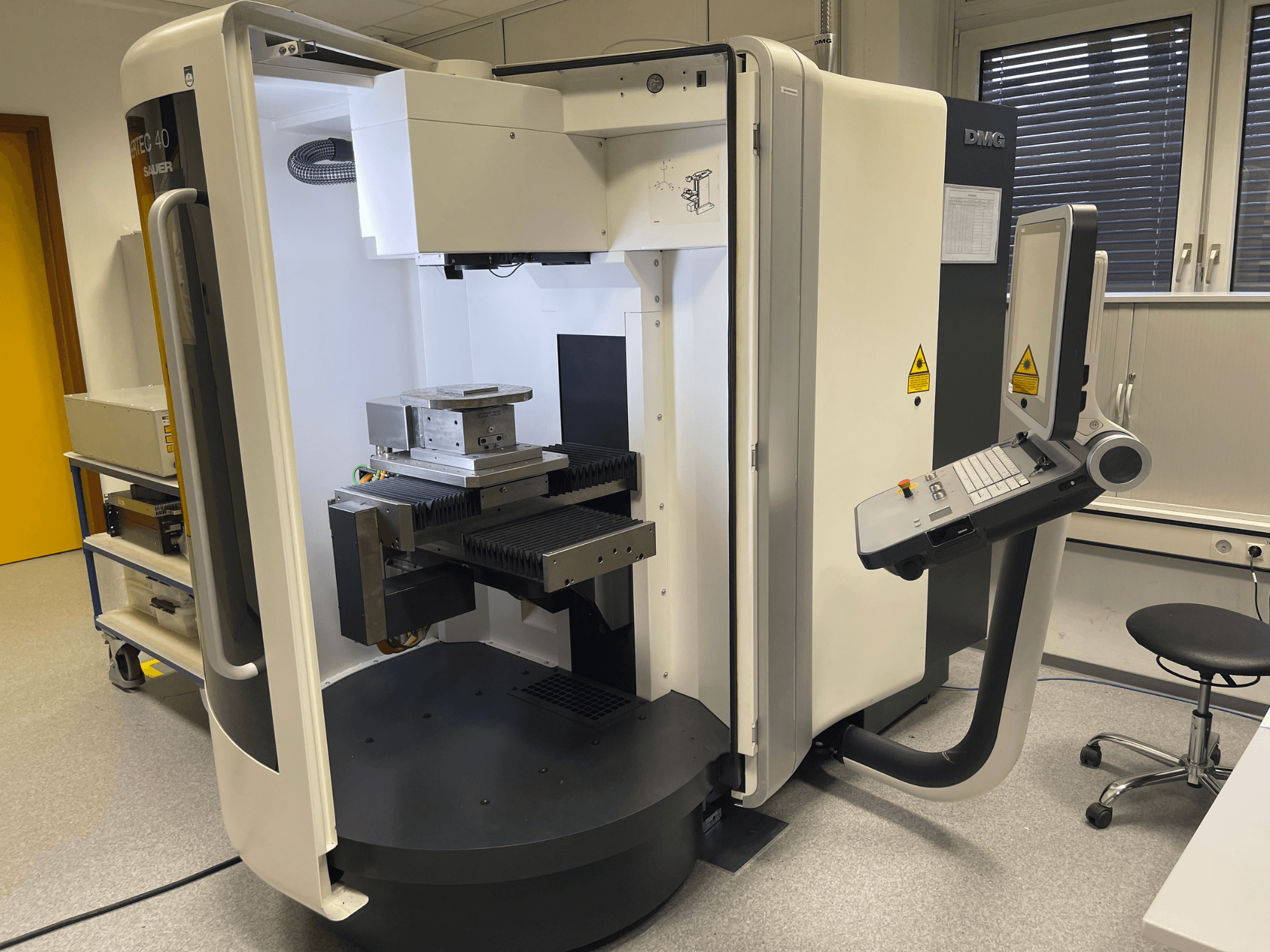 DMC 40 mašīna, skats no sāniem, ar vadības paneli un darba galdu ar metāla detaļu, kas novietota laboratorijas vidē.