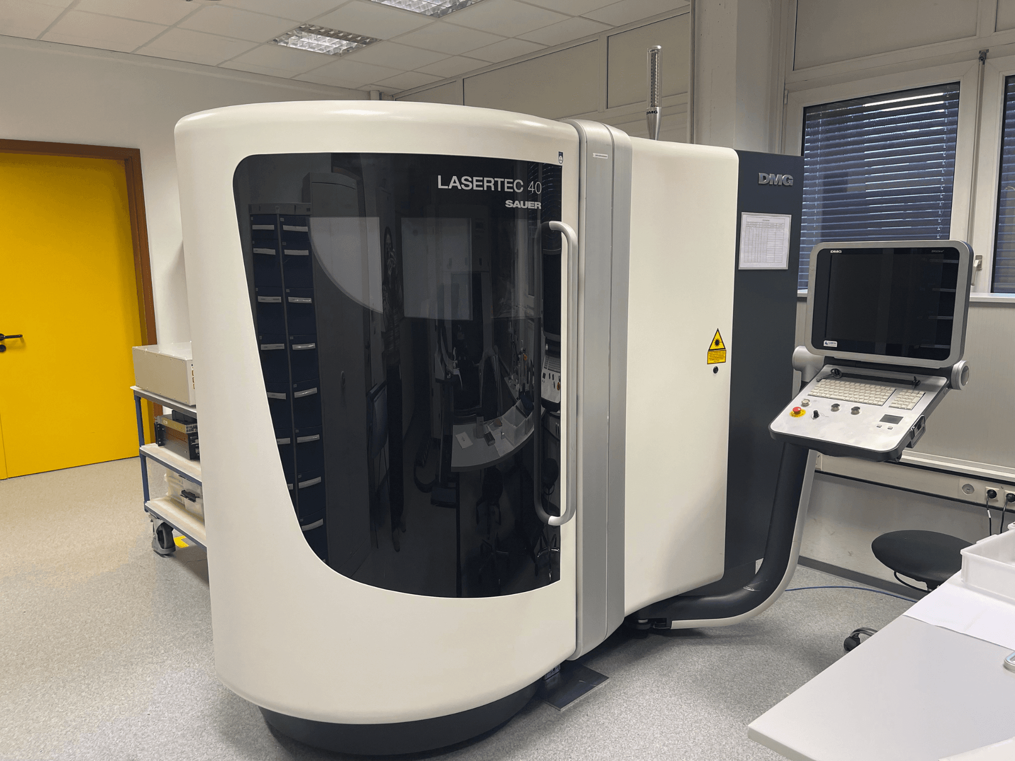 DMG MORI baltā LASERTEC 40 frēzmašīna, skats no priekšpuses, ar vadības paneli un ergonomisku dizainu labi apgaismotā telpā.