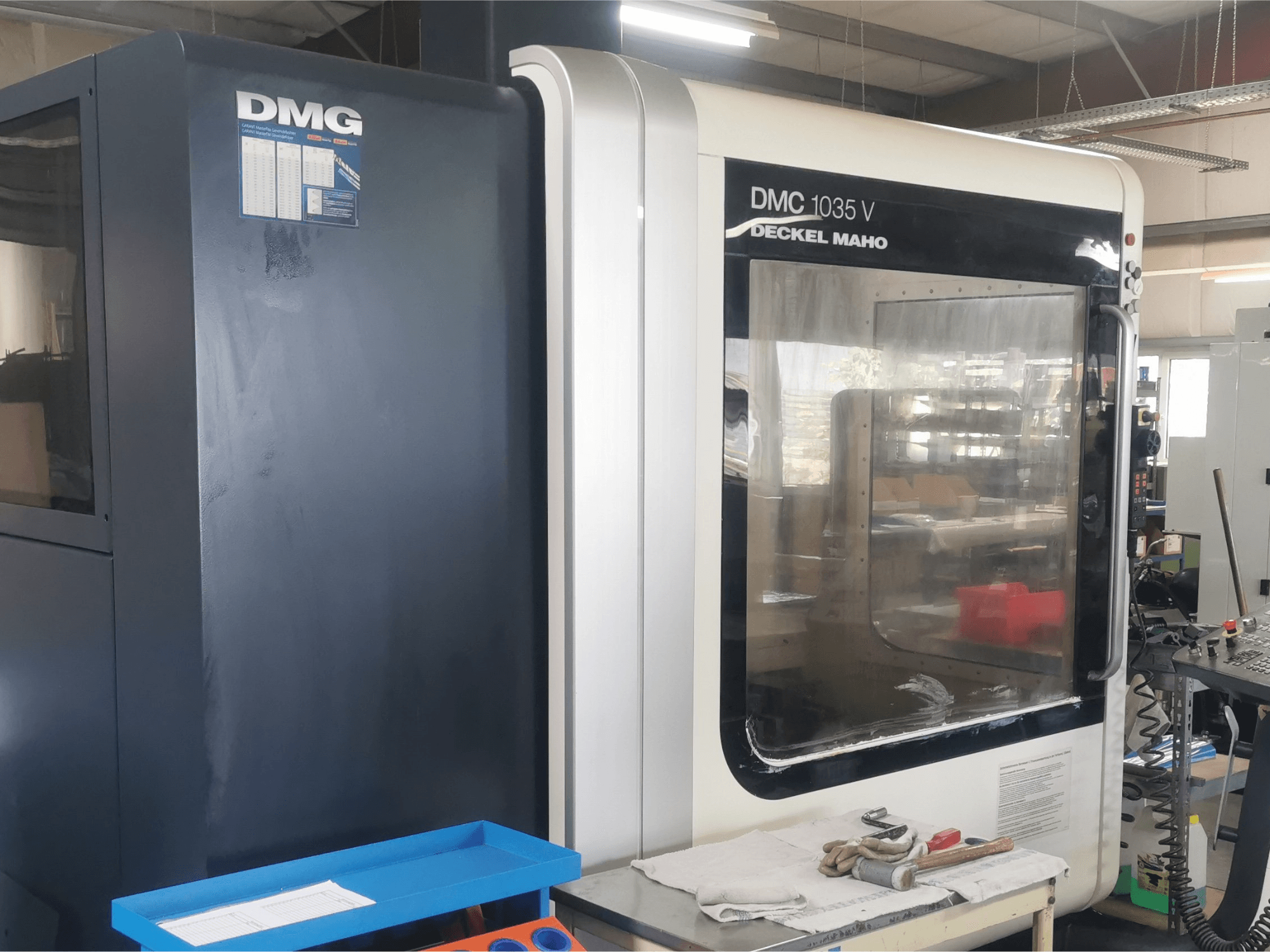 DMG DMC 1035 V frēzmašīna darbnīcā, priekšējais skats ar zilu instrumentu paliktni un cimdiem uz virsmas.
