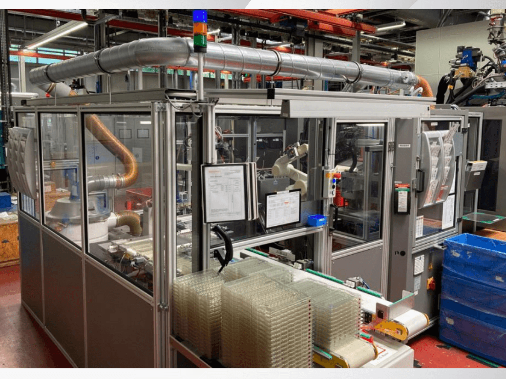 Automatizēta montāžas iekārta ar robotizētu roku, caurspīdīgiem sānu paneļiem un sakrautiem konteineriem ar caurspīdīgiem priekšmetiem, skats no priekšpuses.