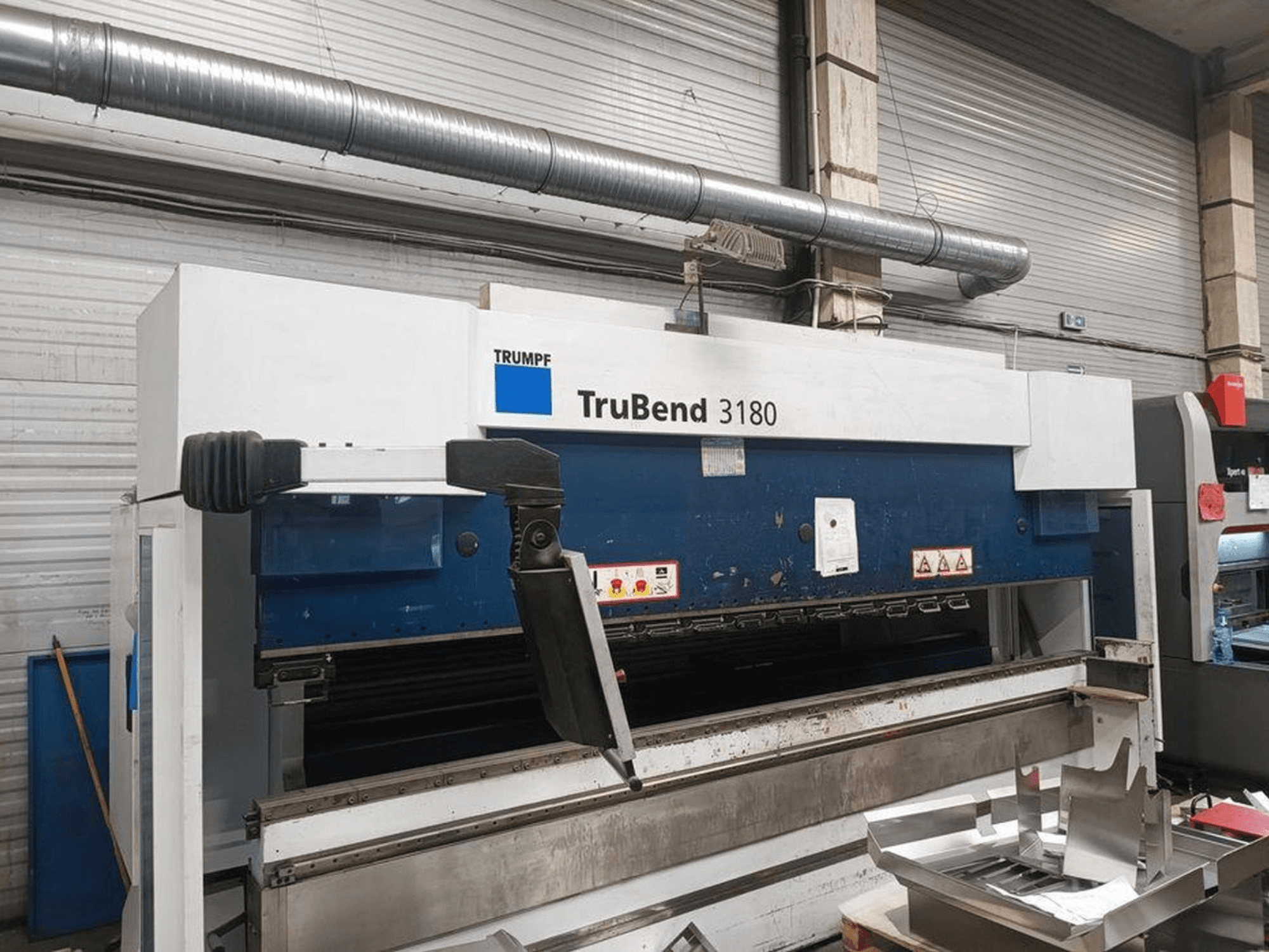 Trumpf TruBend 3180 liekšanas iekārta darbnīcā, sānu skats, kurā redzams vadības panelis un metāla gabali uz galda.