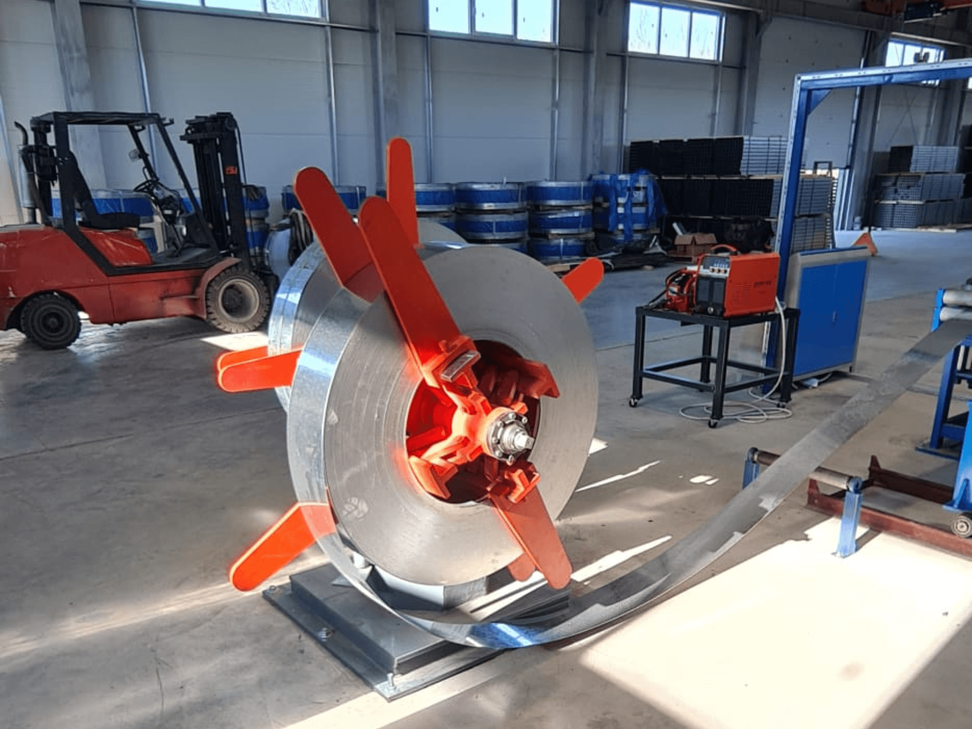 Metāla spole ar sarkaniem rokturiem uz statīva, skatoties no leņķa. Rūpniecisks fons ar mašīnām un iekrāvēju.