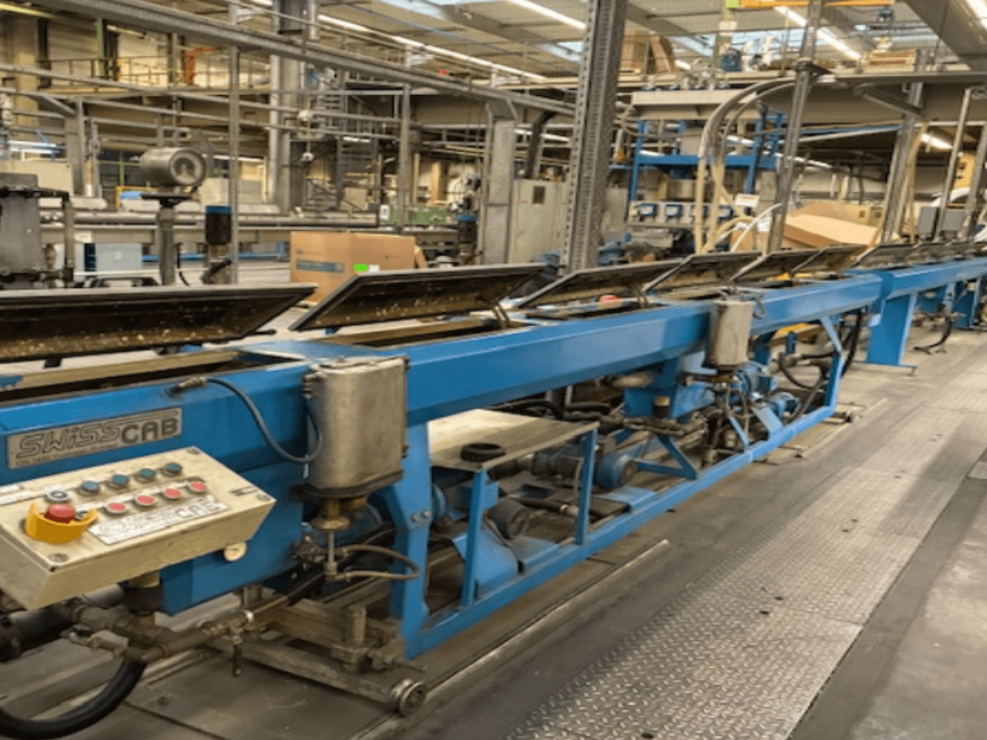 Zilā SwissCab industriālā mašīna rūpnīcā ar vadības pogām un konveijera sistēmu, skats no sāniem.