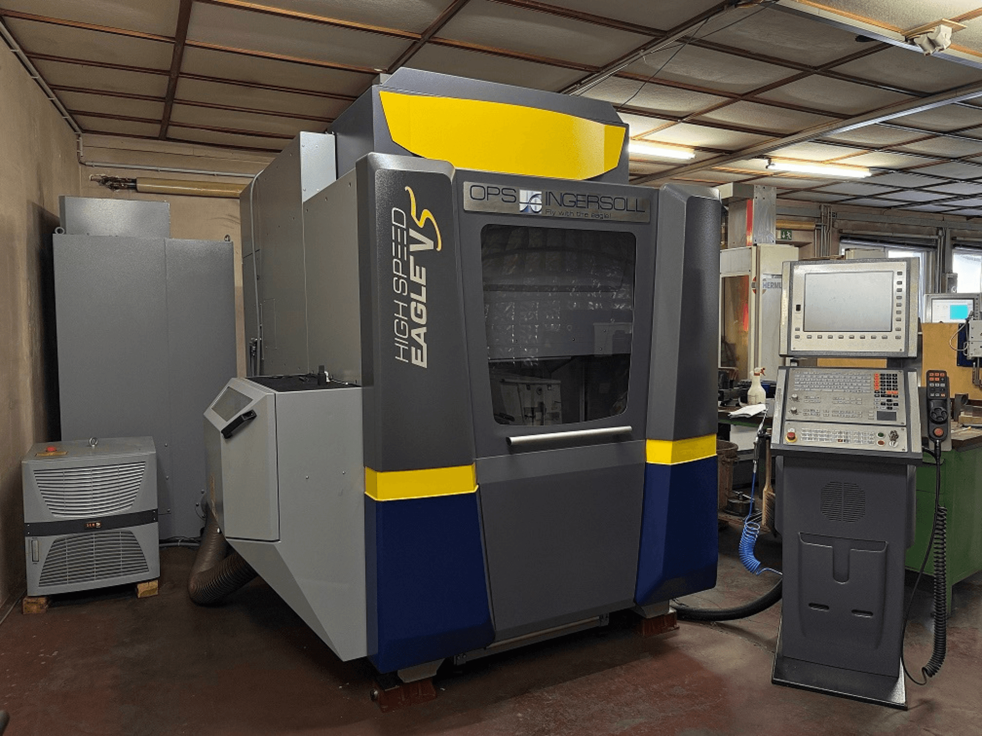 OPS Ingersoll ātrgriezējmašīna Eagle V, skats no sāniem, ar vadības paneli un blakus esošajām iekārtām rūpnieciskā vidē.