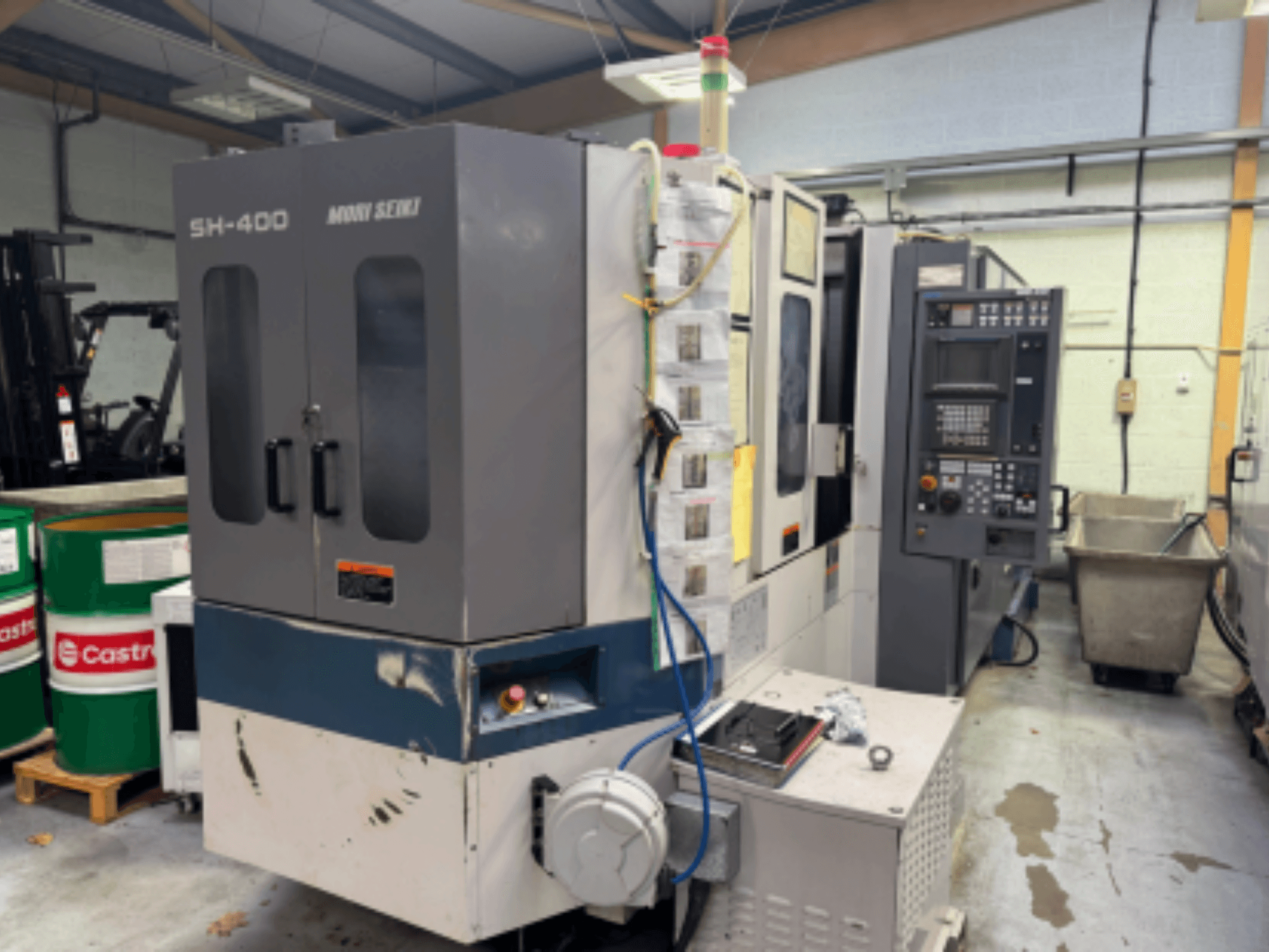 Moni Seiki SH-400 CNC mašīna, sānu skats, kurā redzams vadības panelis, durvis un apkārtējā industriālā vide.