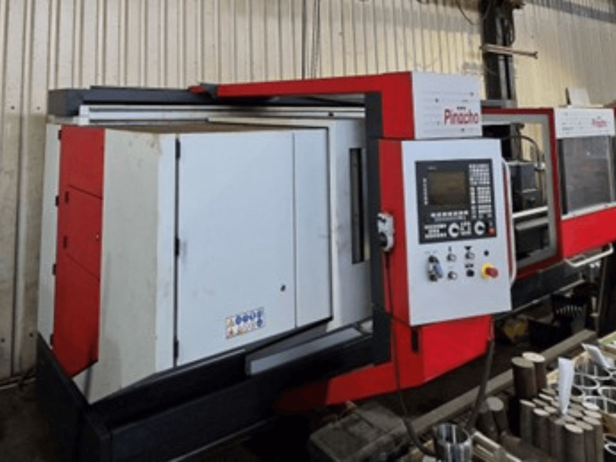 Sarkanbaltā krāsā krāsotas Pinacho CNC darbgaldi, skats no sāniem, ar vadības paneli un materiāliem uz grīdas, parādot darbības iestatījumus.