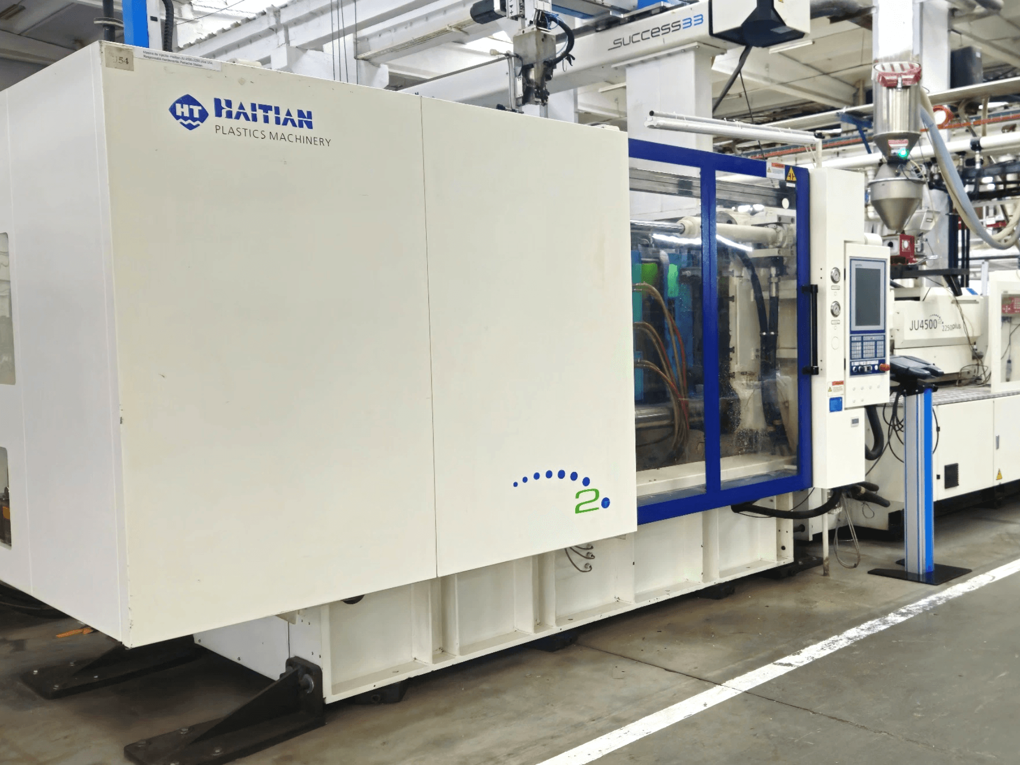 Haitian Plastics Machinery modelis, skats no priekšpuses, kurā redzama mašīna ar vadības paneli un redzamām caurulēm rūpnieciskā vidē.