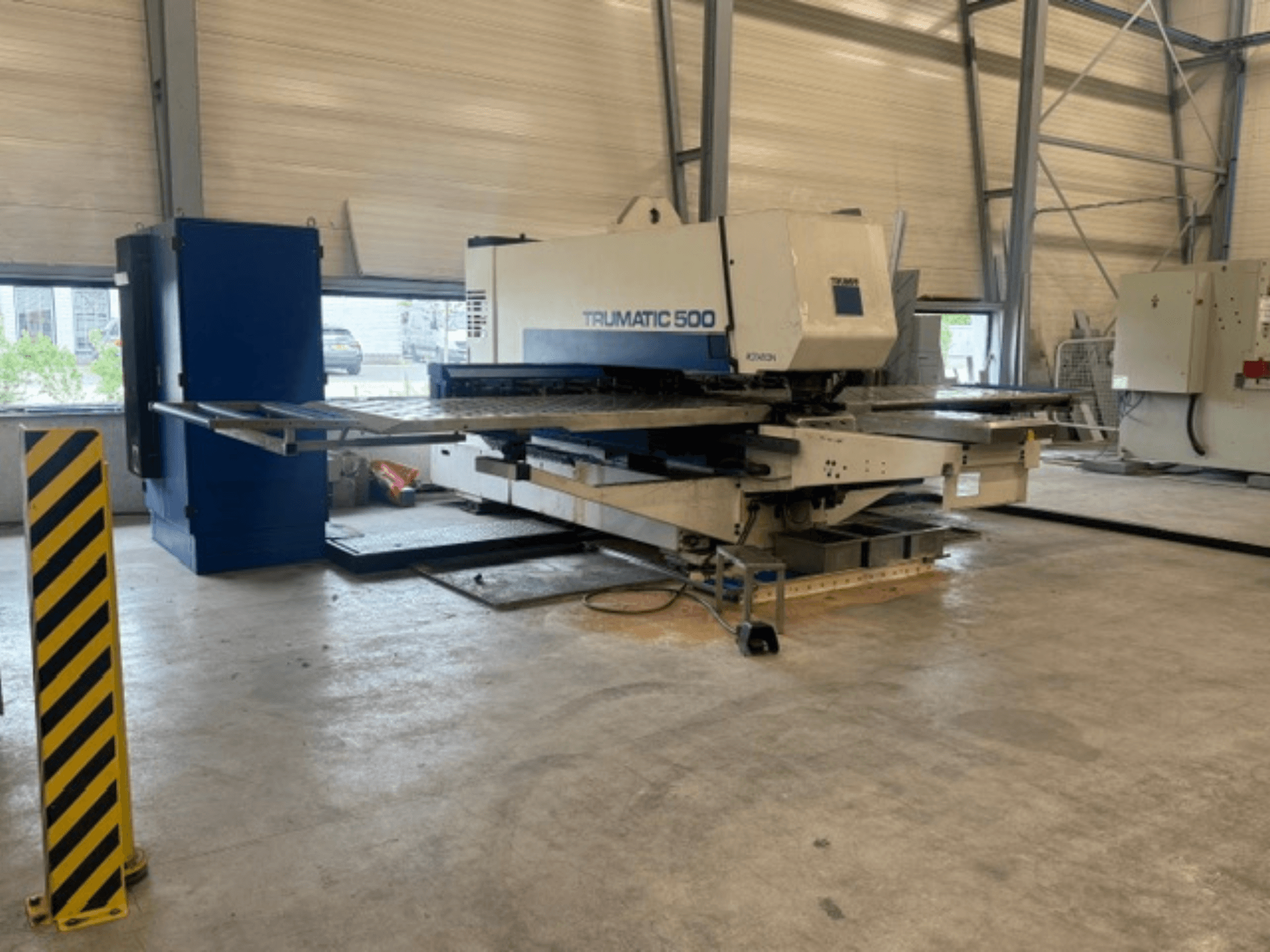 CNC perforācijas mašīna TRUMPF Trumatic 500 darbnīcā, sānskats, kurā redzama iekārta un drošības stabs ar dzeltenām svītrām.
