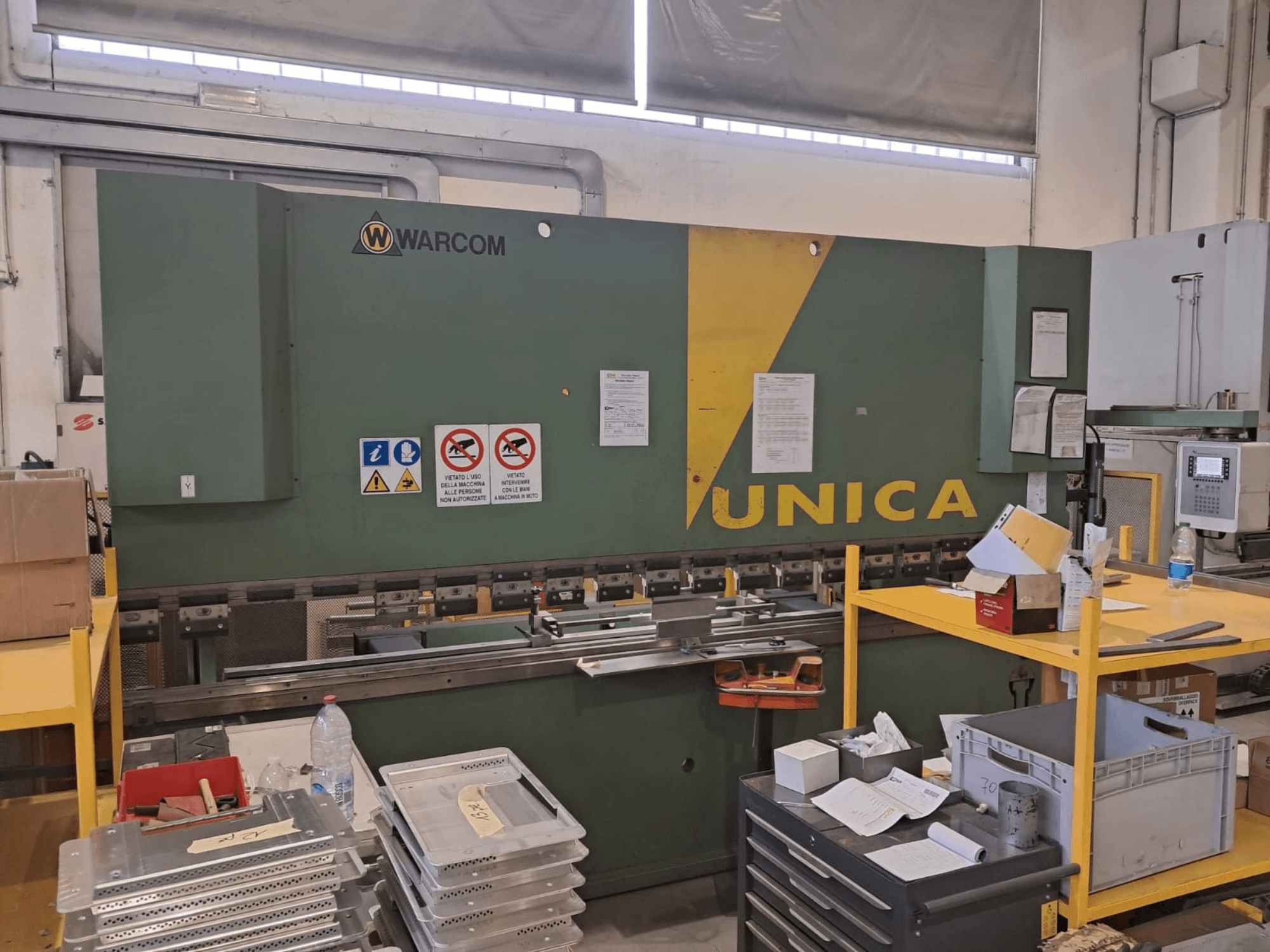 Hidrauliskā prese „Warcom UNICA 100-40”, skats no priekšpuses, zaļš korpuss ar dzelteniem akcentiem, redzamas drošības zīmes, uz galda atrodas darbarīki un materiāli.