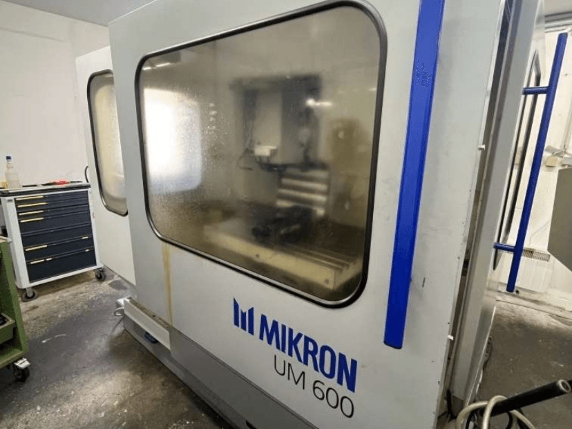 MIKRON UM 600 TNC 410/426 universālais apstrādes centrs, skats no sāniem, ar miglotiem stikliem un redzamu instrumentu uzglabāšanas vietu blakus.