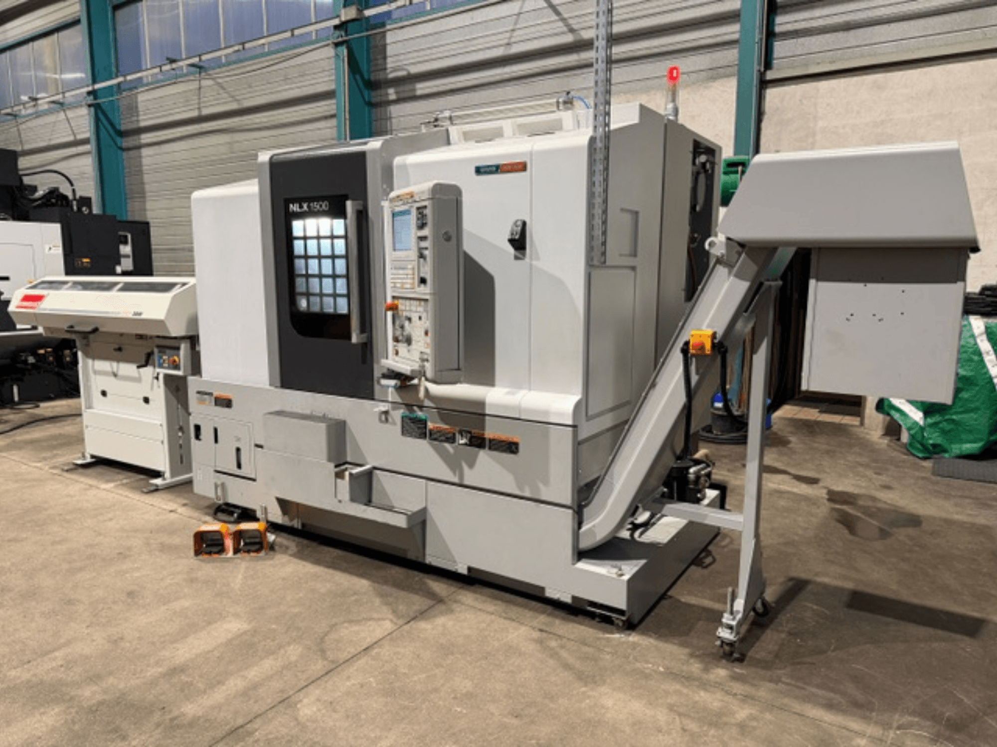NLK 1500 CNC mašīna, skats no priekšpuses, ar vadības paneli un piederumu tvertni sānos, novietota uz betona grīdas.