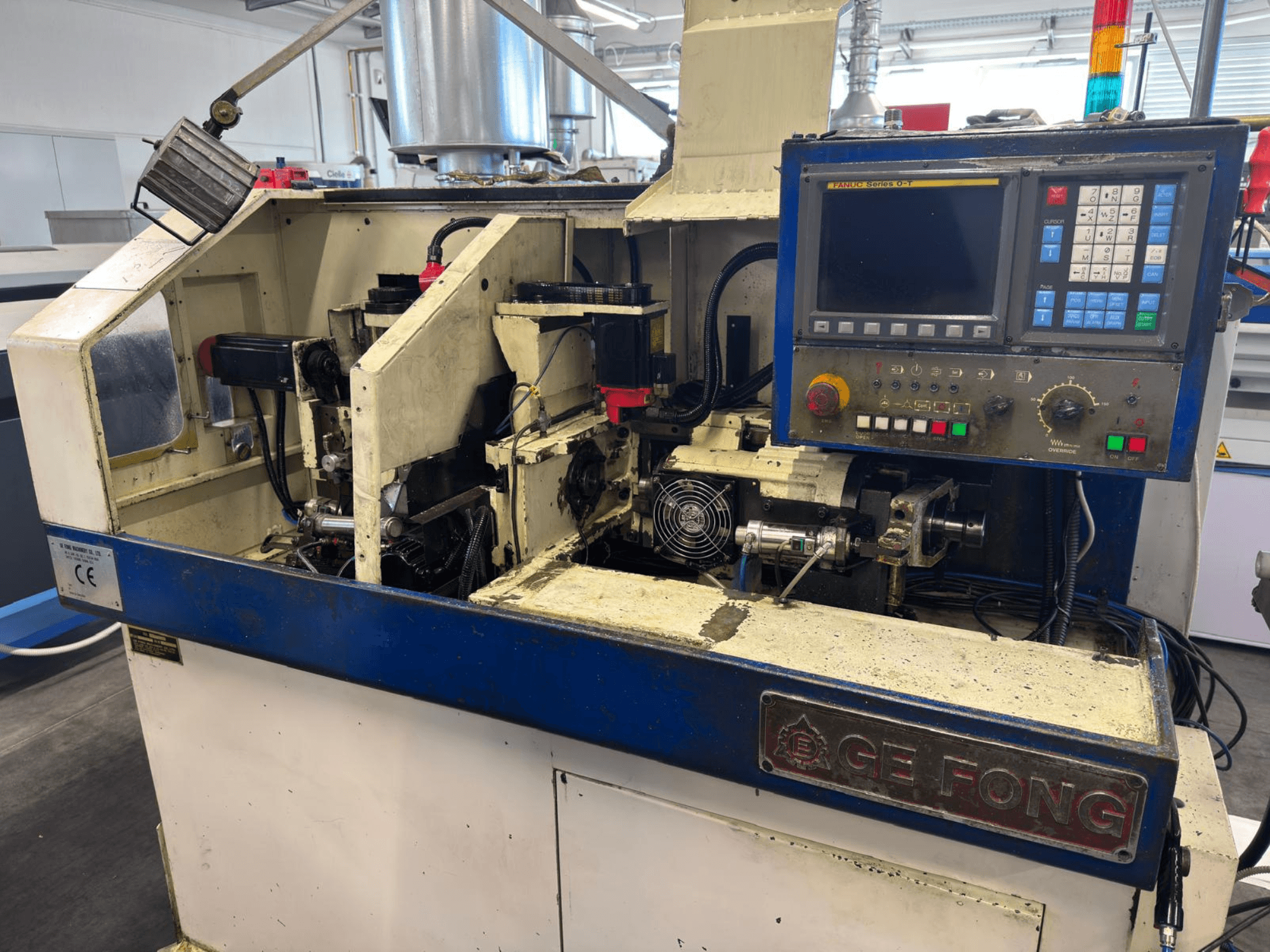 GE FONG industriālā mašīna priekšpusē, kurā redzami iekšējie komponenti, vadības panelis un darba detaļas.