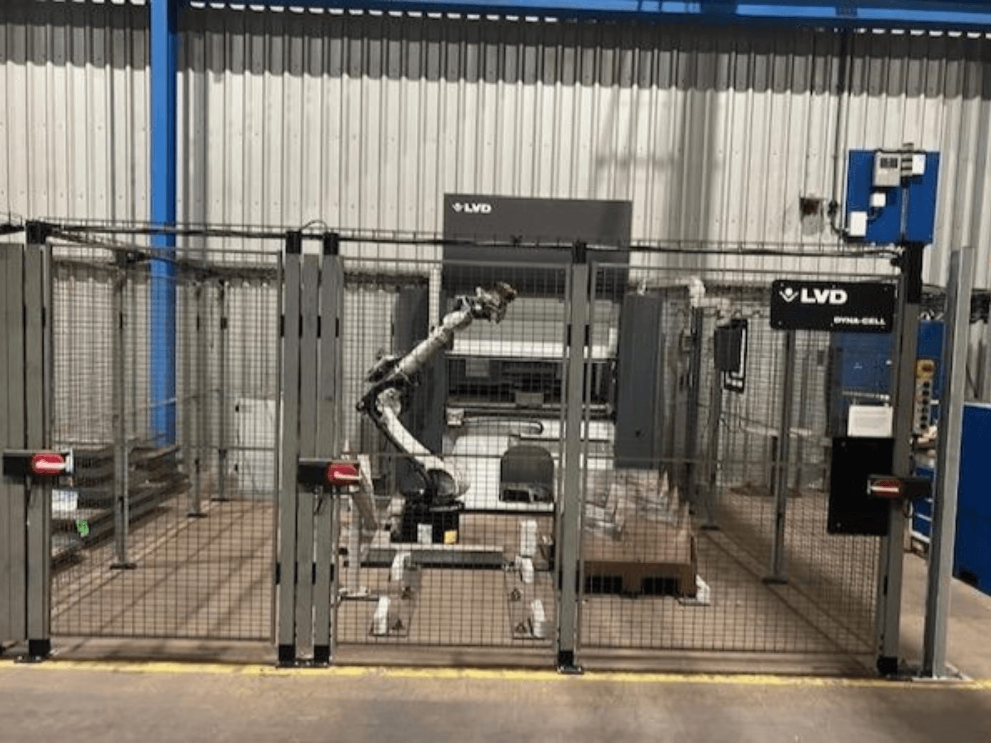 LVD Dyna-Cell robotizētā darbstacija iežogotā korpusā, skats no priekšpuses, ar robotizēto roku un vadības paneli.