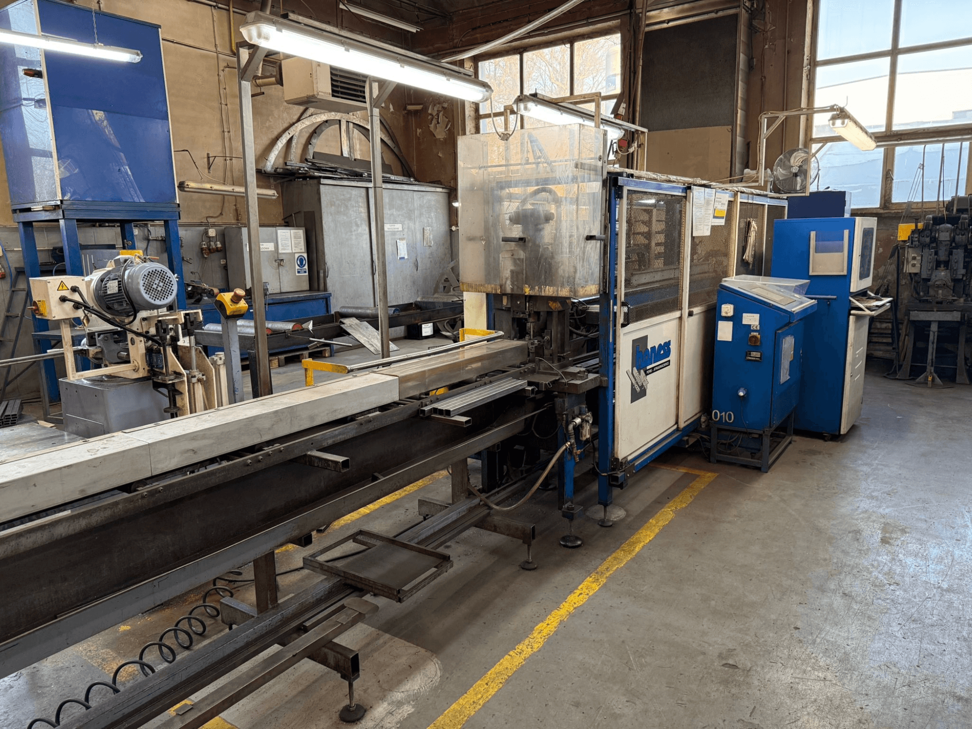 Rūpnieciskā darbnīca ar zilu Haweas mašīnu, skats no sāniem, kurā redzams konveijers un iekārtas lielā, labi apgaismotā telpā.