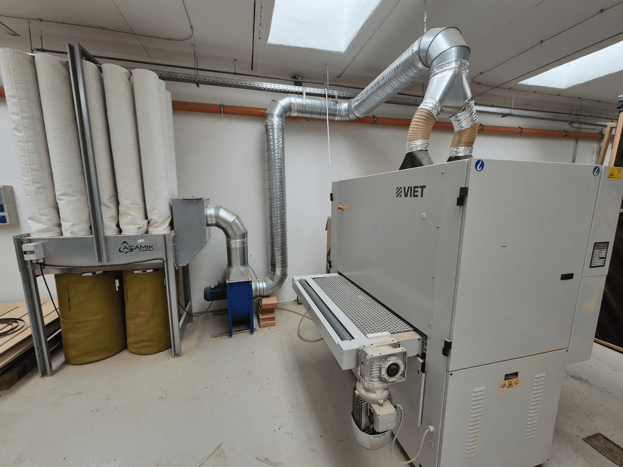 Viet putekļu nosūces sistēma ar cilindriskiem filtriem un sudraba izplūdes sistēmu, skatoties no priekšpuses darbnīcā.