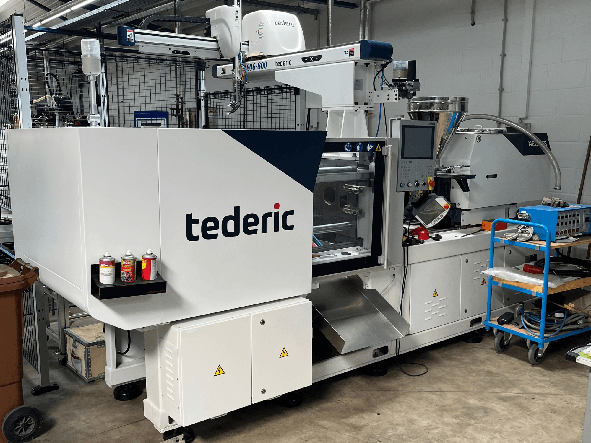 Hidrauliskā injekcijas formēšanas mašīna TEDERIC NEO.E55/e110h darbnīcā, skats no sāniem ar vadības pulti un blakus esošajiem instrumentiem.