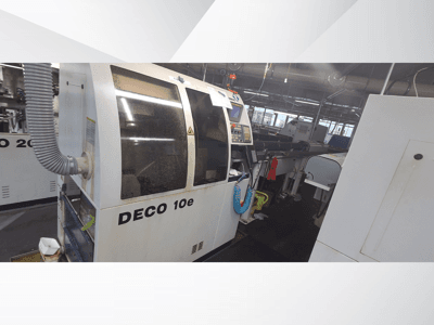 DECO 10e mašīna darbnīcā; skats no sāniem, kurā redzams vadības panelis un ventilācijas šļūtene, fonā redzamas neskaidras iekārtas.
