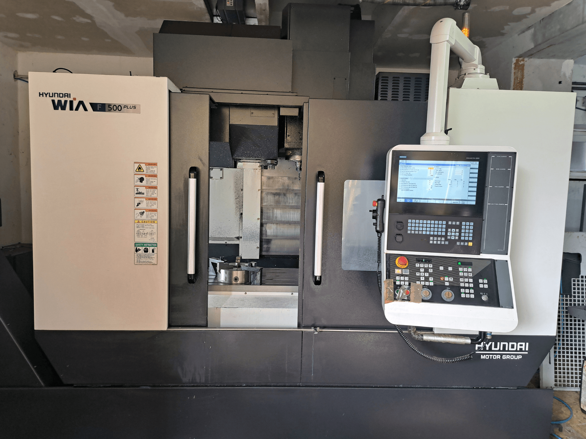 Hyundai WIA F500 Plus CNC mašīna priekšpusē ar digitālo vadības paneli un drošības marķējumu.