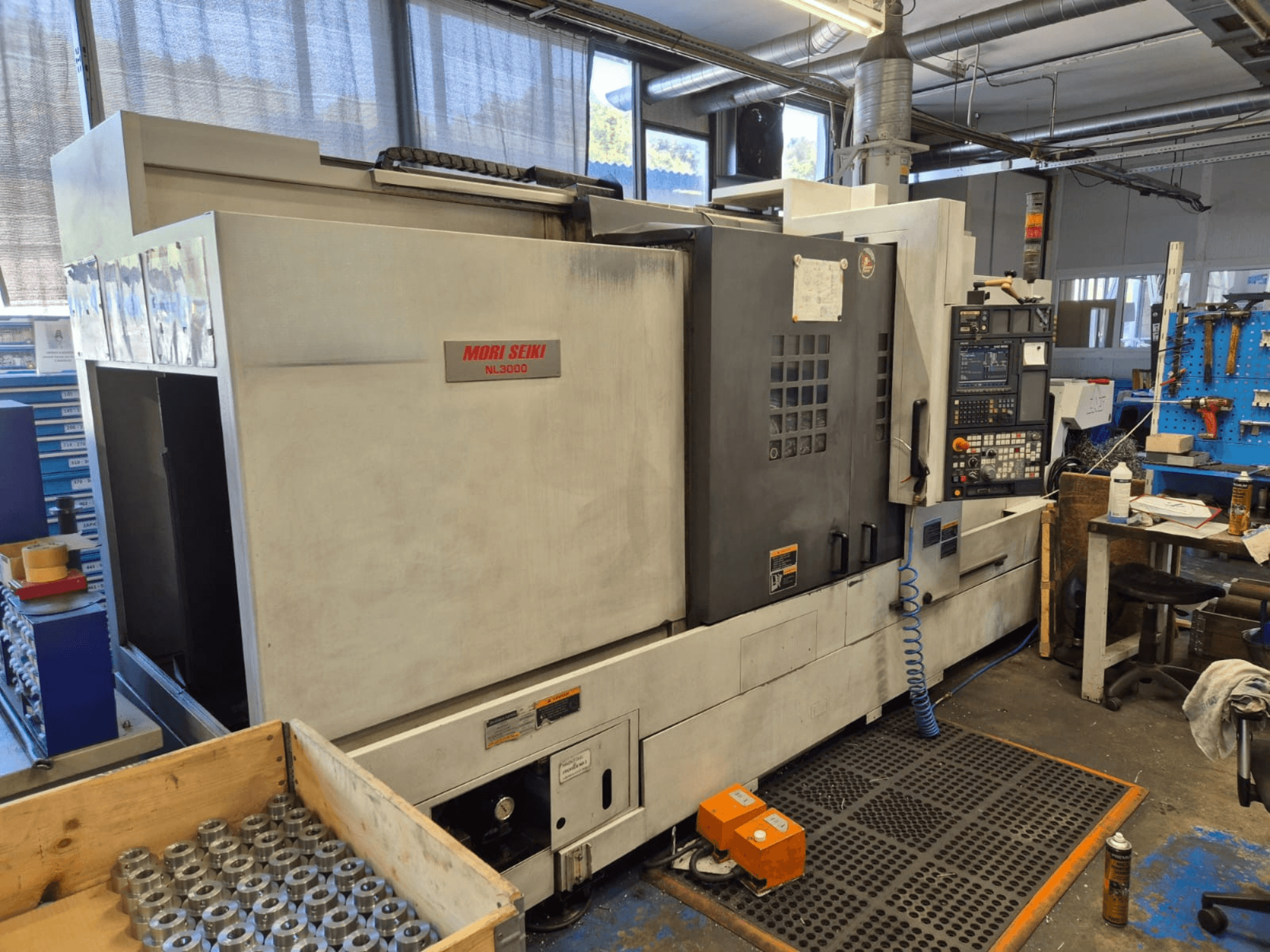 Mori Seiki NL3000 CNC virpa darbnīcā, sānu skats, kurā redzams vadības panelis, instrumenti un metāla detaļas koka kastē.