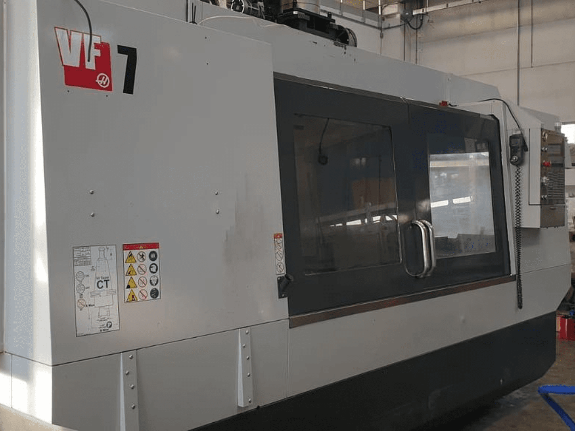 Haas VF-7 CNC darbgalds sānu skatā, ar metāla pelēku apdari, vadības paneli un drošības marķējumu.